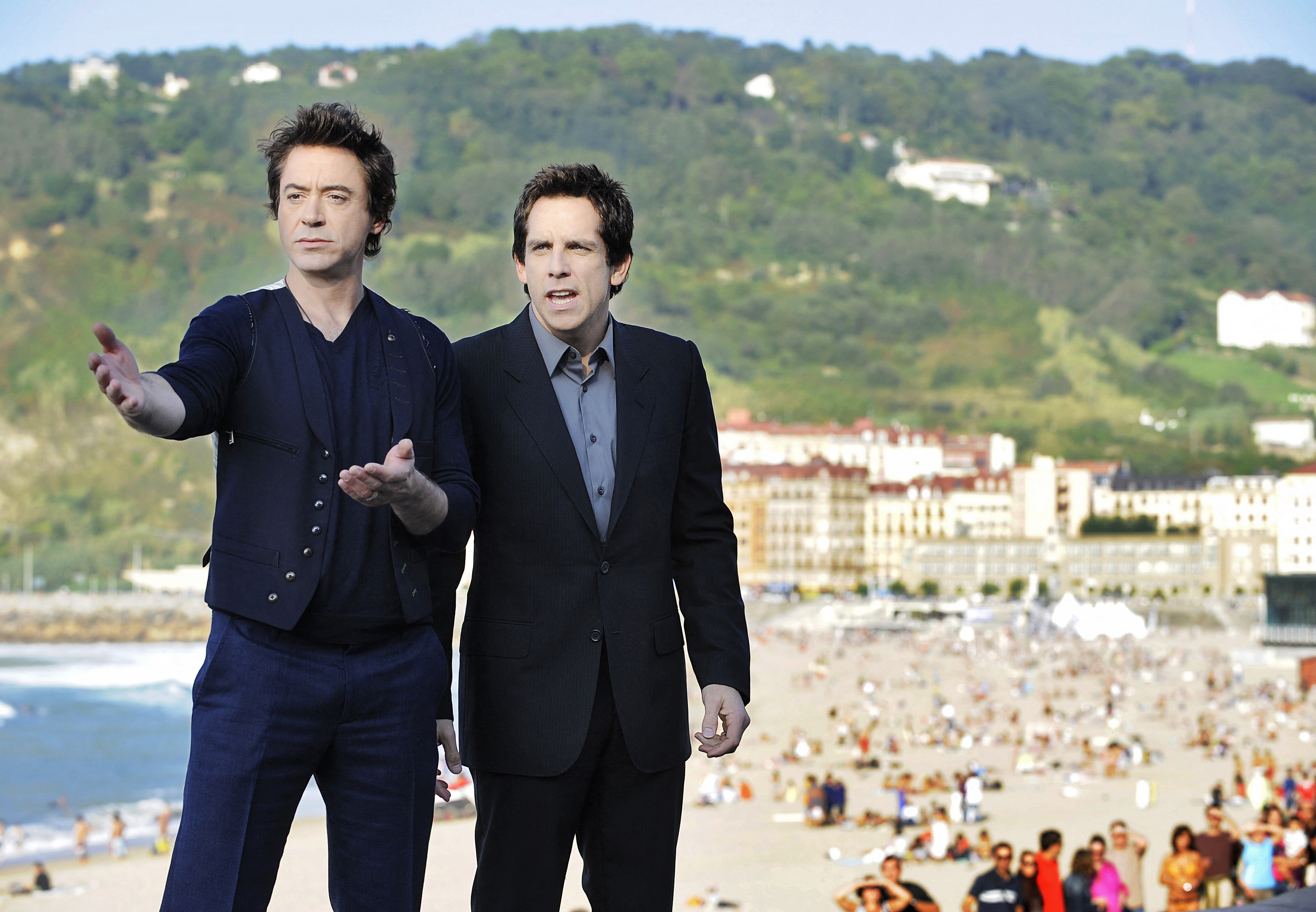 Two men in dark suits stand outdoors above a crowded beach with a hillside and grand hotel building in the background.