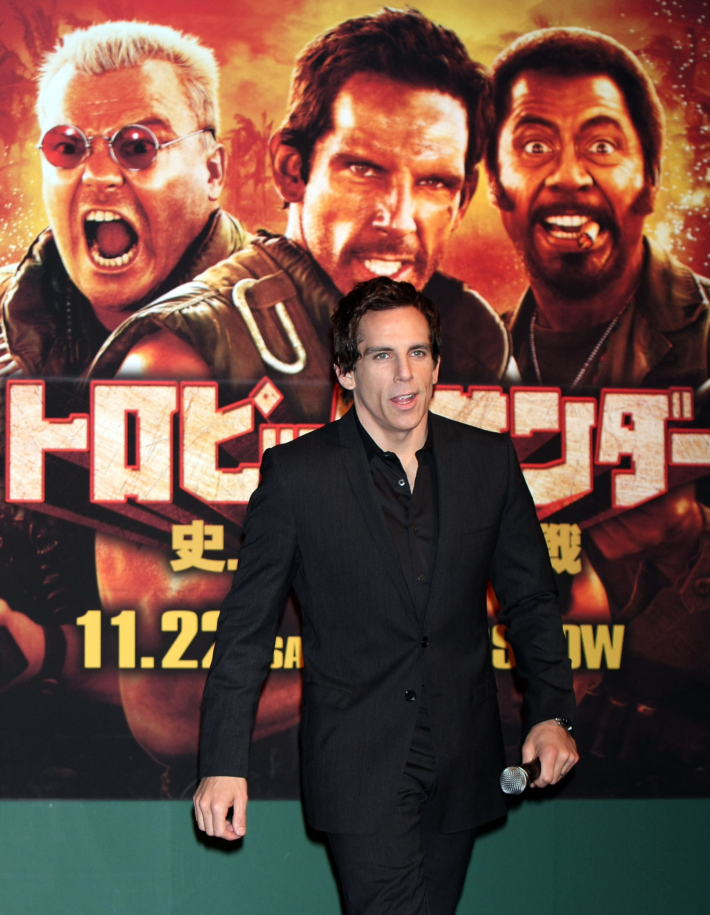 Ben Stiller in a black suit holding a microphone, standing in front of a Japanese Tropic Thunder movie poster at a Tokyo press conference.