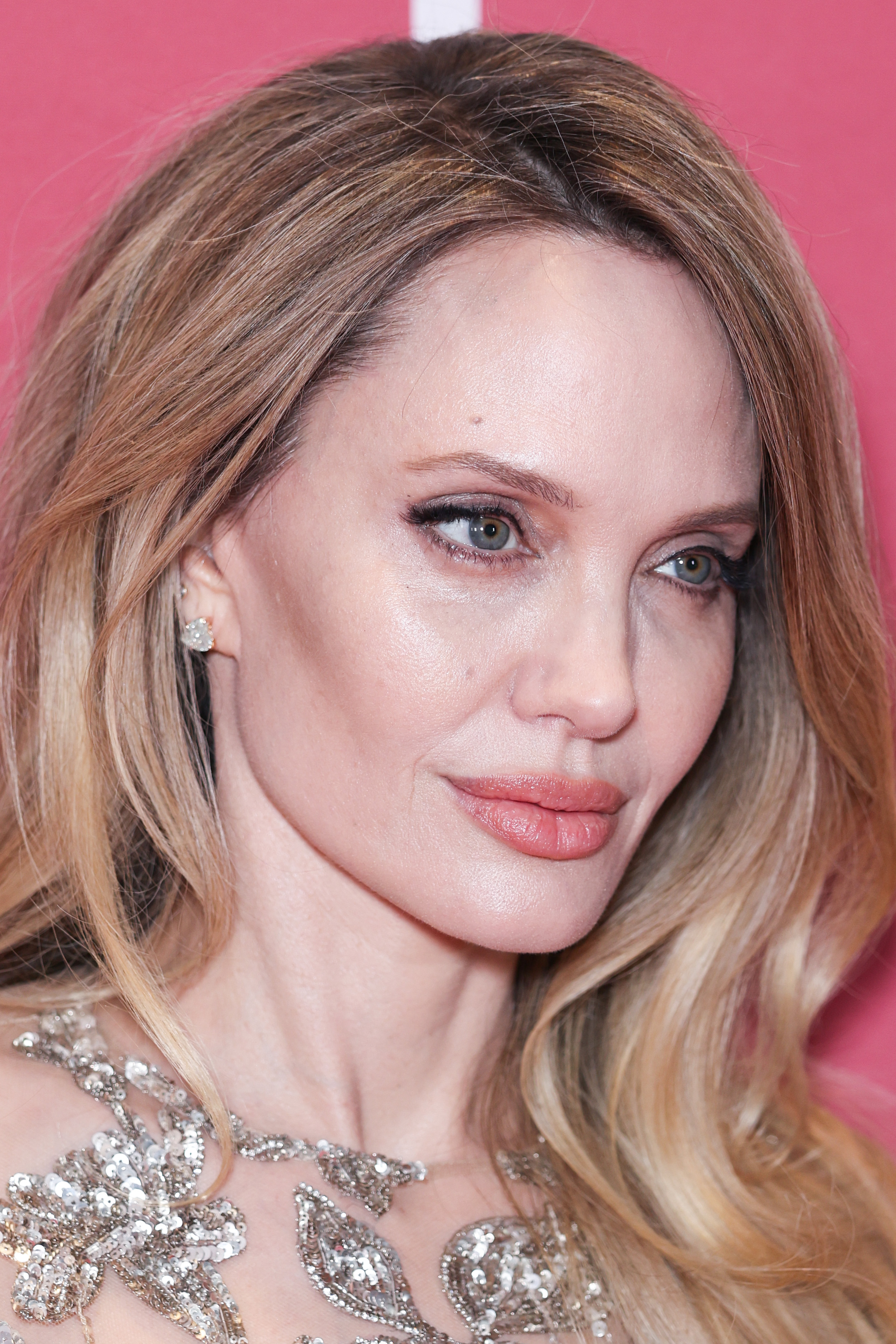 Close-up of a woman with long blonde hair wearing a silver sequined dress, posing in front of a pink backdrop.