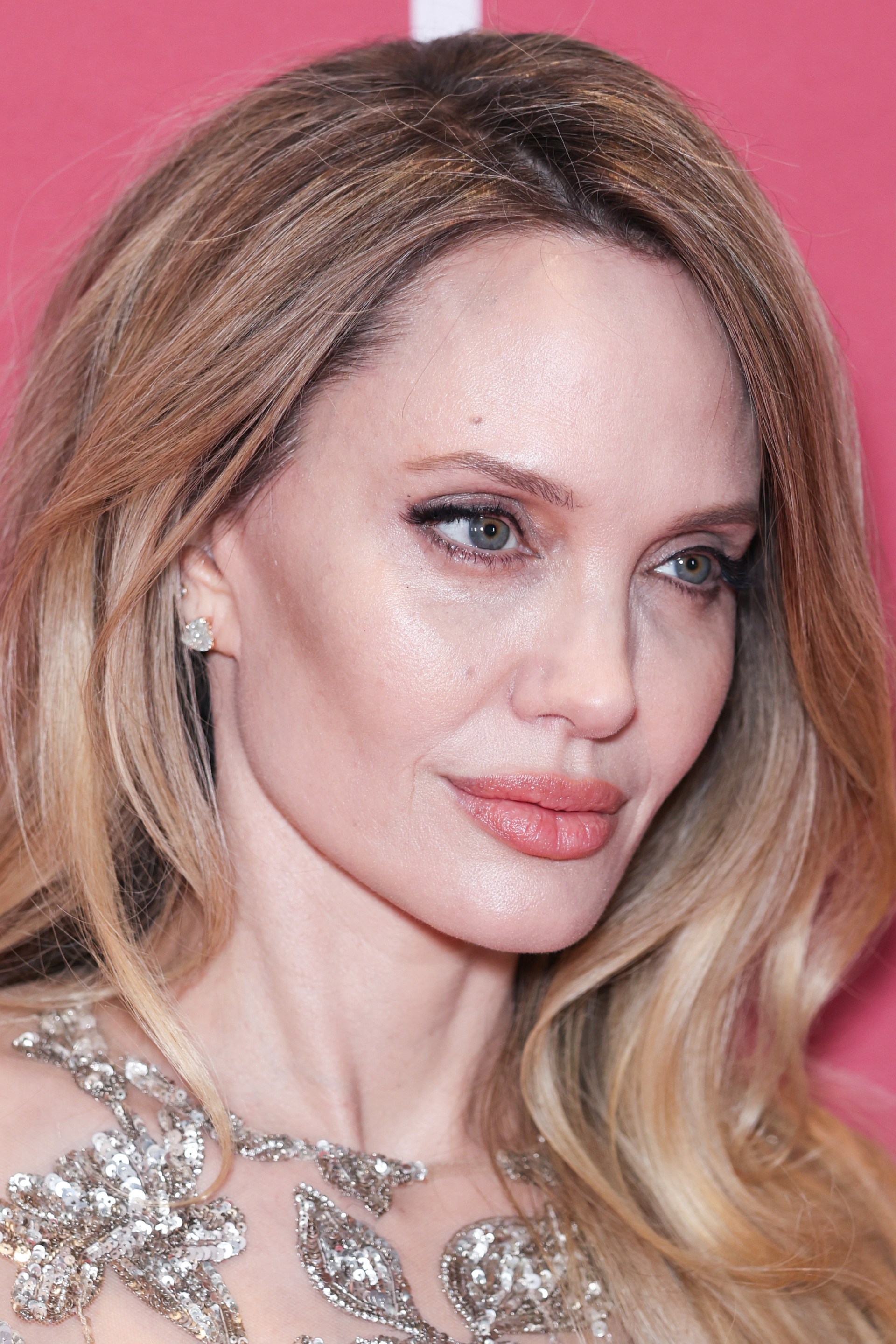 Close-up of a woman with long blonde hair wearing a silver sequined dress, posing in front of a pink backdrop.