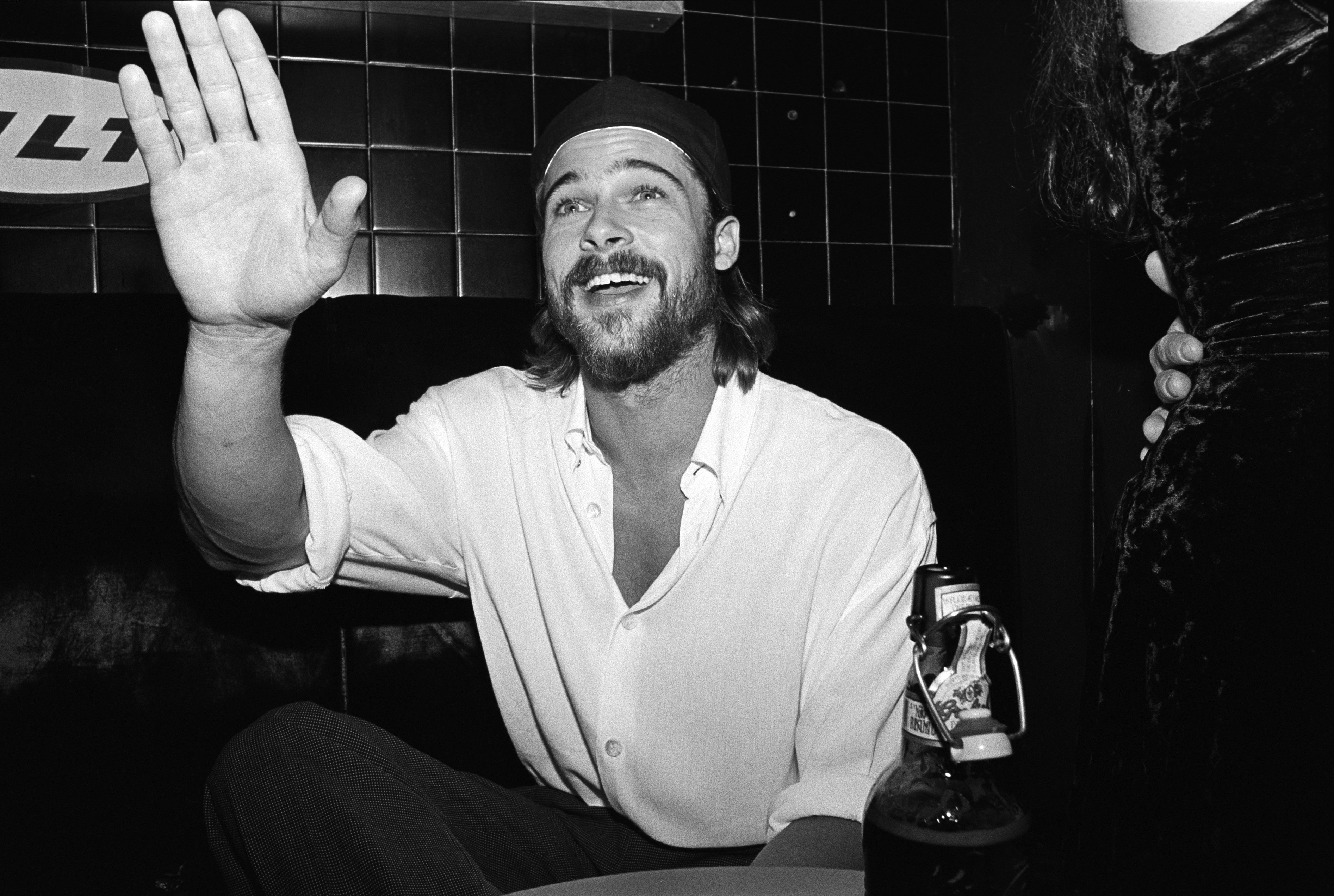 Black and white photo of a young man with a beard and knit cap waving and smiling at a bar or club, holding a bottle.