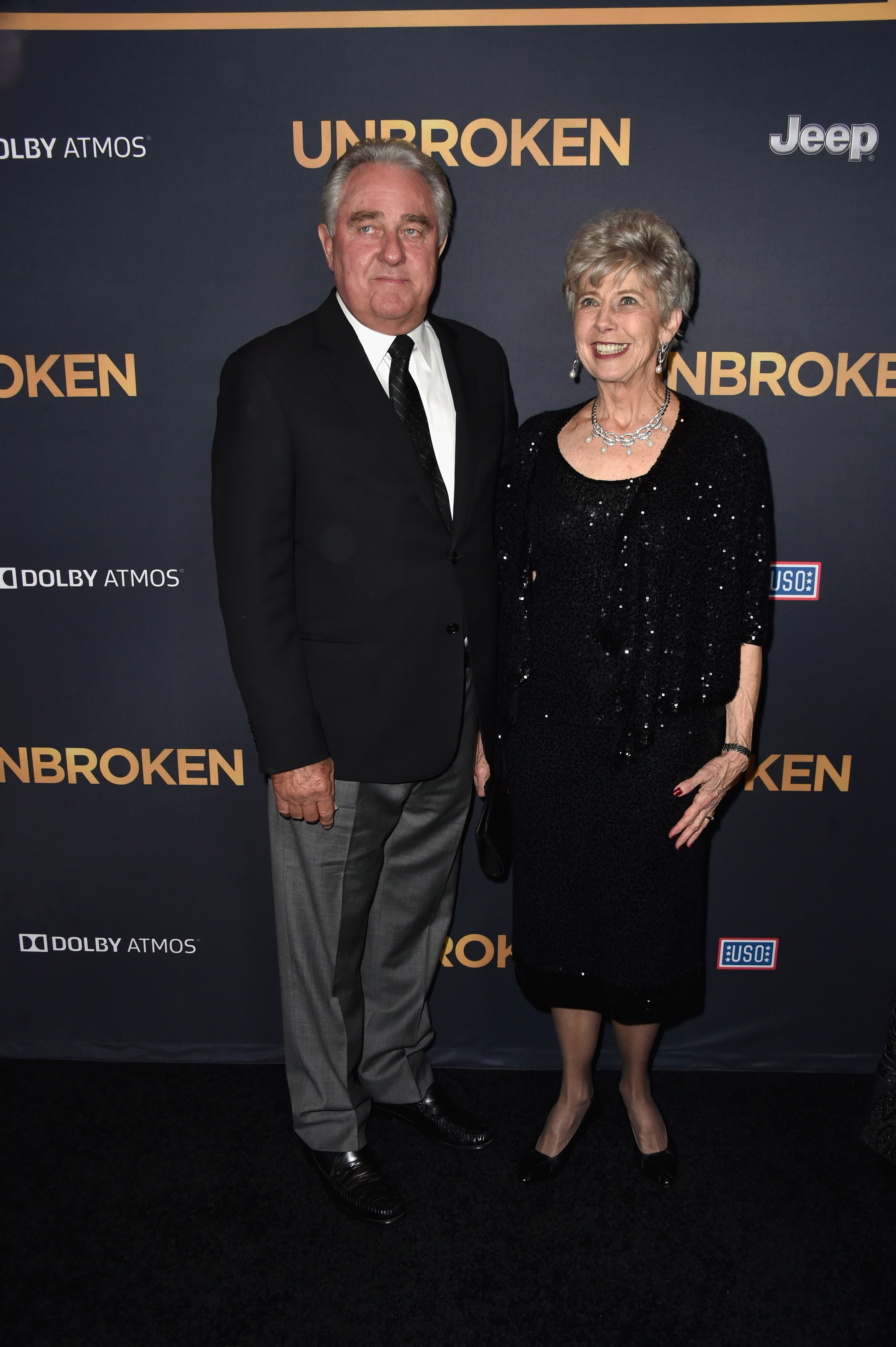 William Pitt in a black suit and Jane Pitt in a black sequined dress standing in front of an Unbroken movie premiere backdrop.