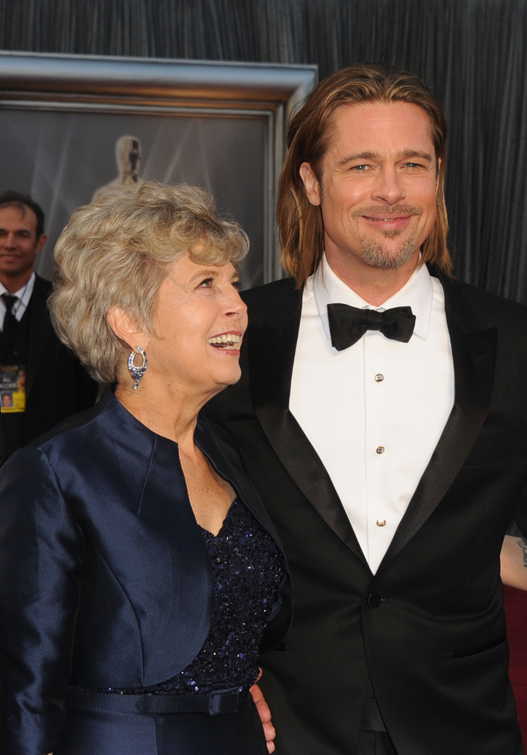 Brad Pitt in a tuxedo and his mother Jane Pitt in a navy gown smiling together on the Oscars red carpet.