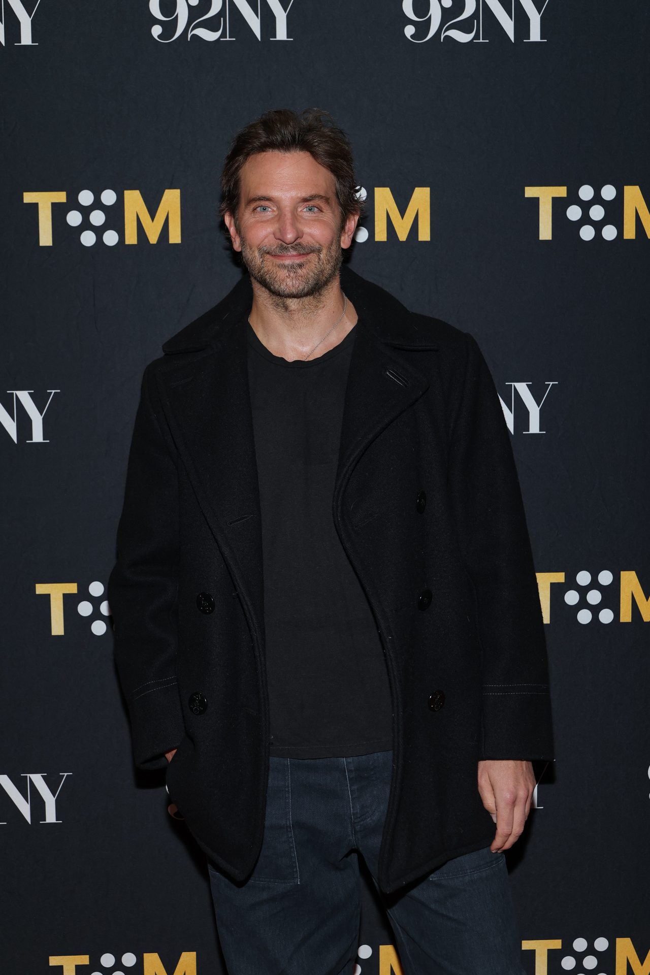 Bradley Cooper smiling in a black peacoat and jeans in front of a TCM and 92NY step-and-repeat backdrop.
