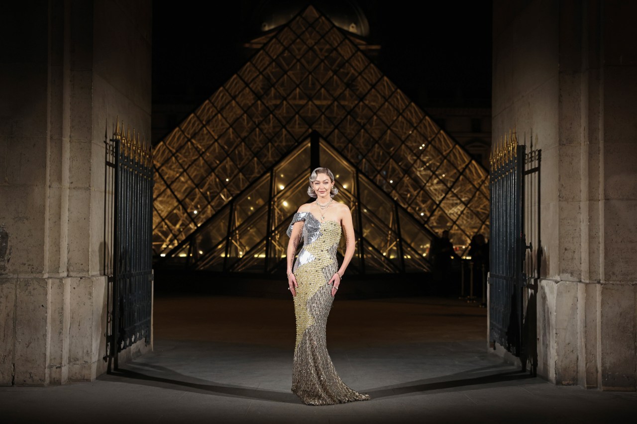 Woman in a silver and gold sequined gown standing in front of the illuminated Louvre Pyramid at night in Paris.