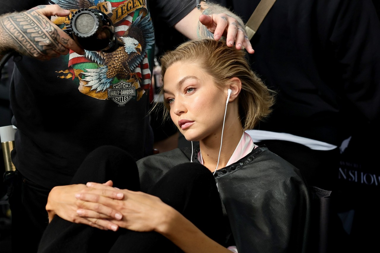 A model in a styling cape with earbuds in sits in a chair while a tattooed hairstylist styles her hair backstage at a fashion show.