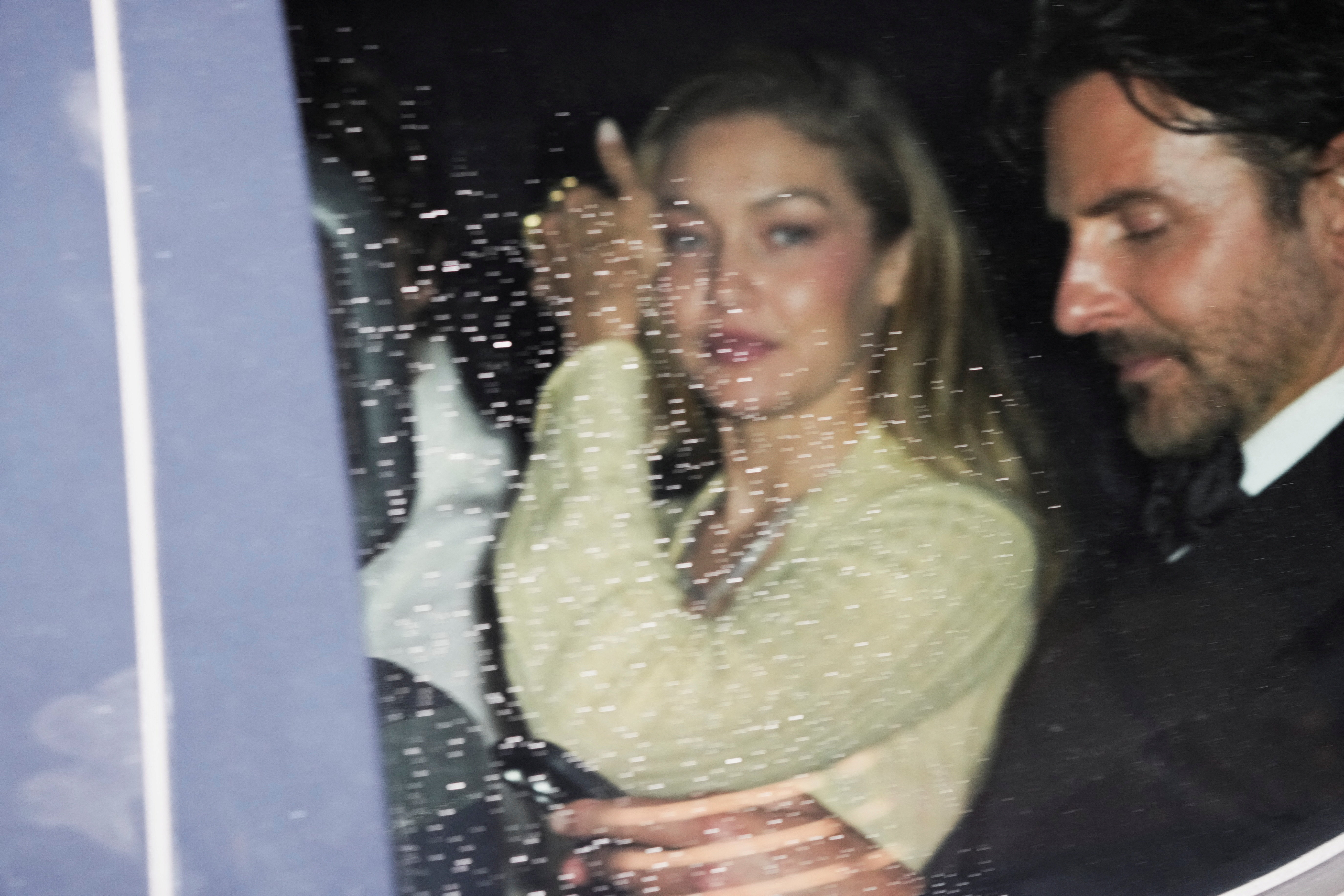 Click to open the image gallery A woman in a cream sweater waves from inside a car next to a dark-haired man in a suit, photographed through a rain-speckled window at night.