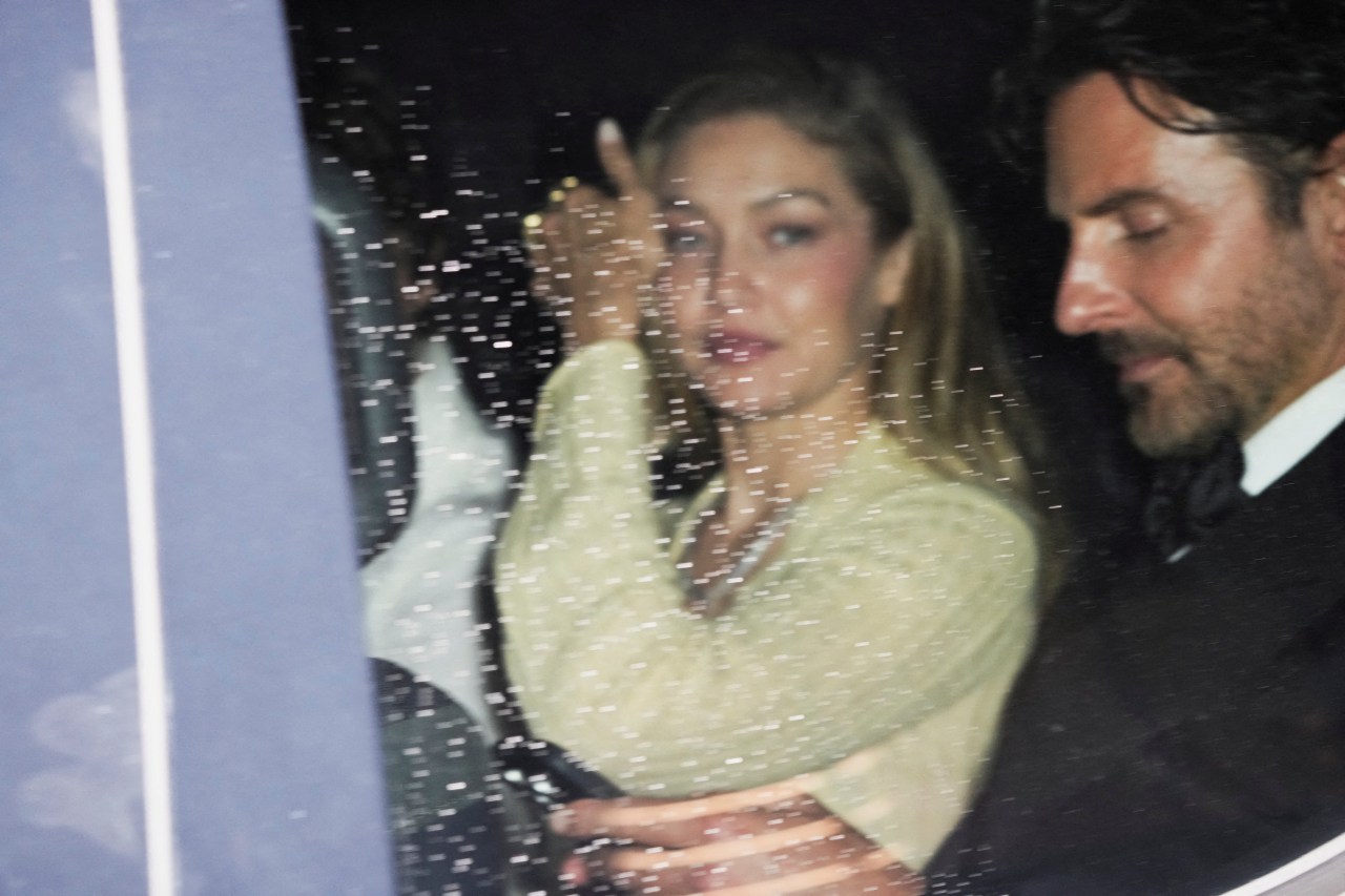 A woman in a cream sweater waves from inside a car next to a dark-haired man in a suit, photographed through a rain-speckled window at night.