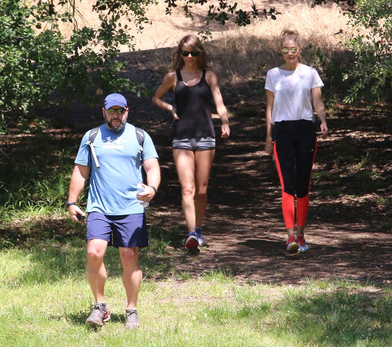 Two women in athletic wear hiking down a dirt trail in a park, accompanied by a man in a blue cap and backpack.