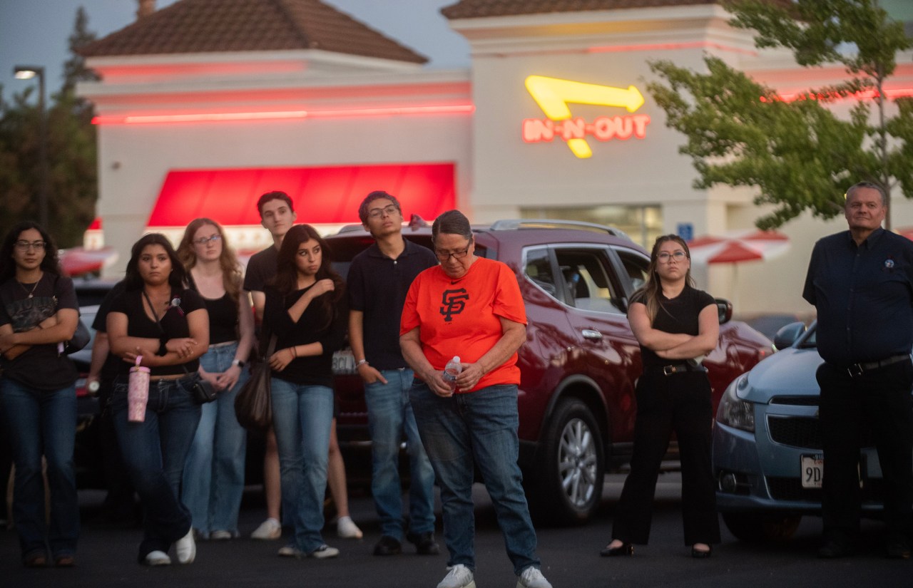 A group of people stand solemnly in a parking lot in front of an In-N-Out Burger at dusk; a woman in an orange SF Giants shirt stands in front with her head bowed.