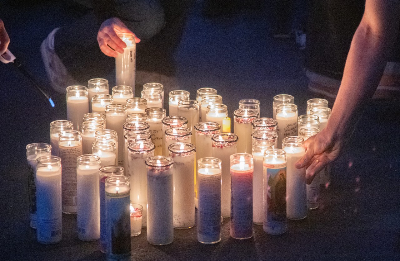 Dozens of lit memorial candles arranged on dark pavement, with two people's hands placing and lighting candles among them.
