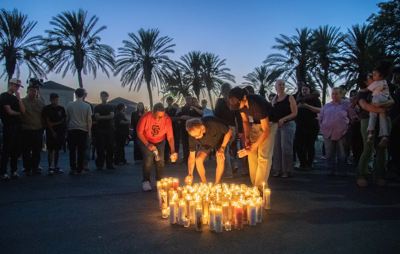 Crowd of people surrounding dozens of lit candles on pavement at dusk, with palm trees silhouetted against a twilight sky.