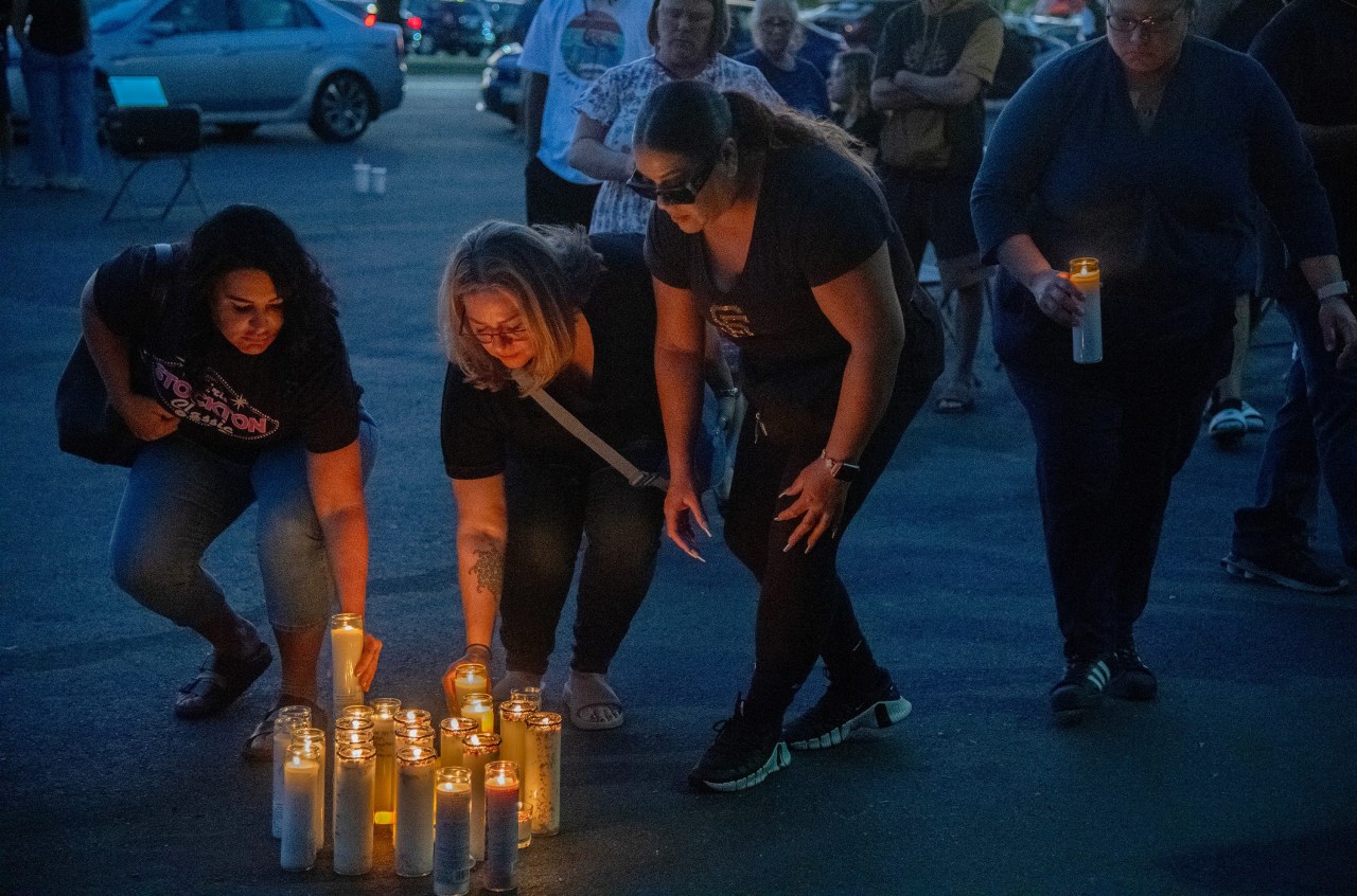 Three women crouch to place lit candles on a parking lot pavement at a candlelight vigil at night, with a crowd gathered behind them.