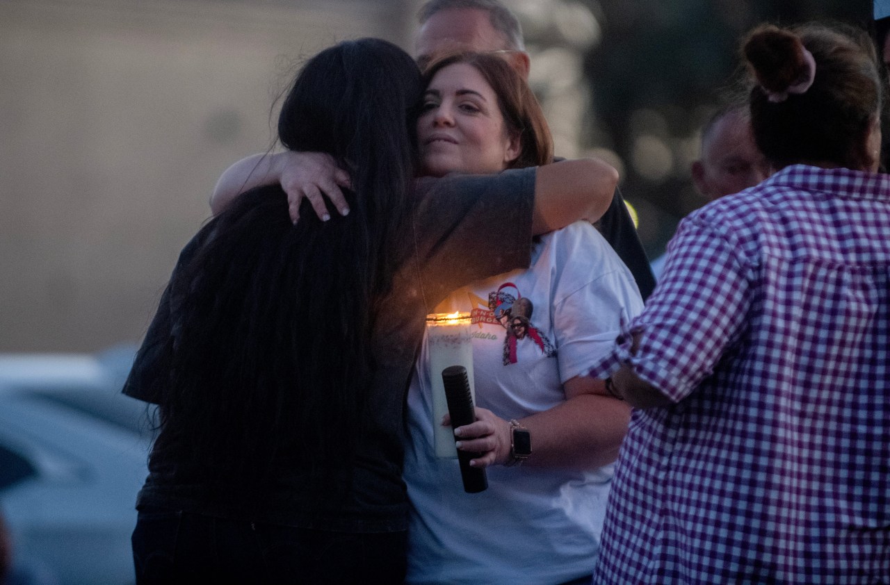 A woman in a white T-shirt holding a candle is hugged by another woman during an outdoor candlelight vigil at night.