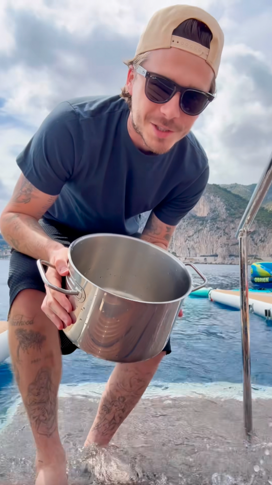 Tattooed man in sunglasses and a backwards cap smiling and holding a large stainless steel pot on the back of a yacht.