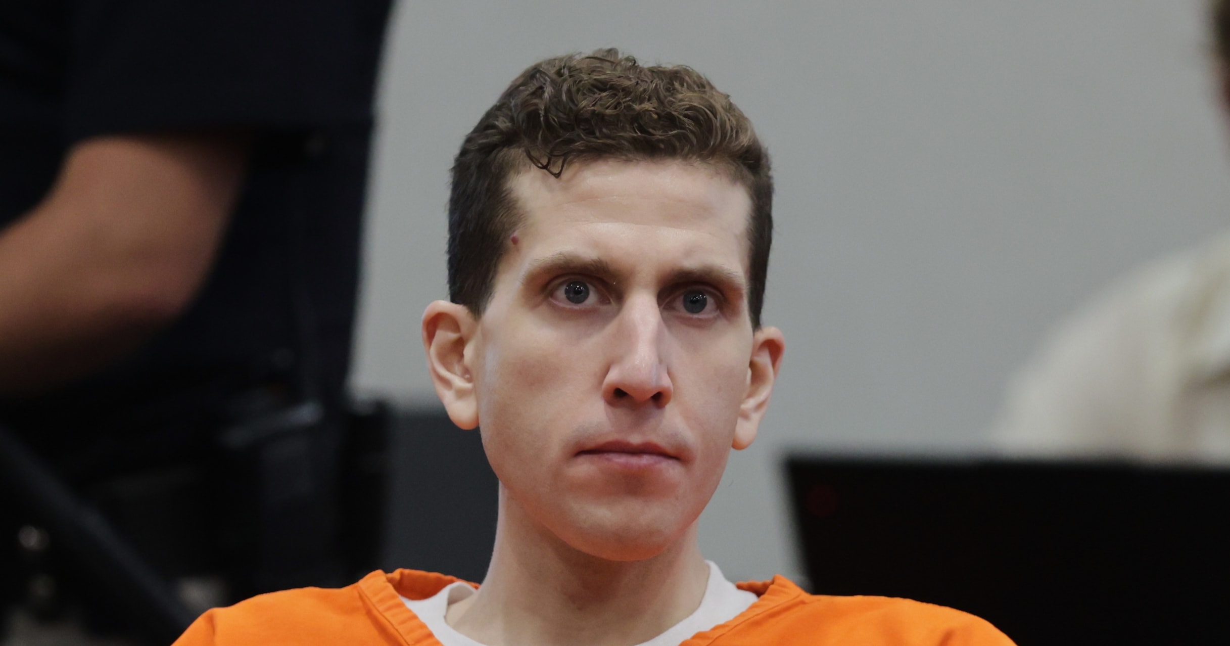 Bryan Kohberger in an orange jail jumpsuit seated at a courthouse sentencing hearing, looking forward with a neutral expression.
