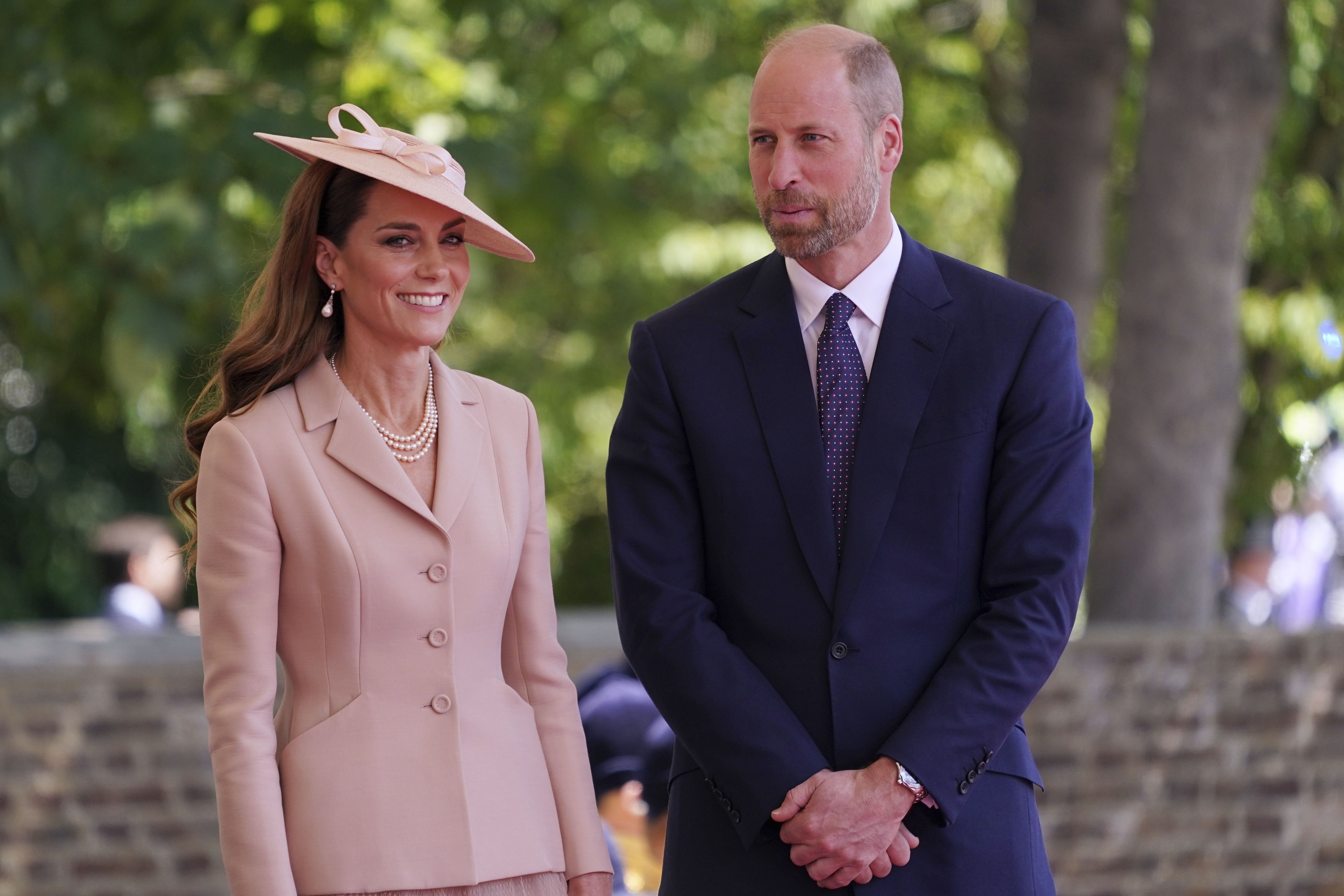 Princess of Wales in a pink suit and hat smiling next to Prince William in a navy suit at Windsor Castle.