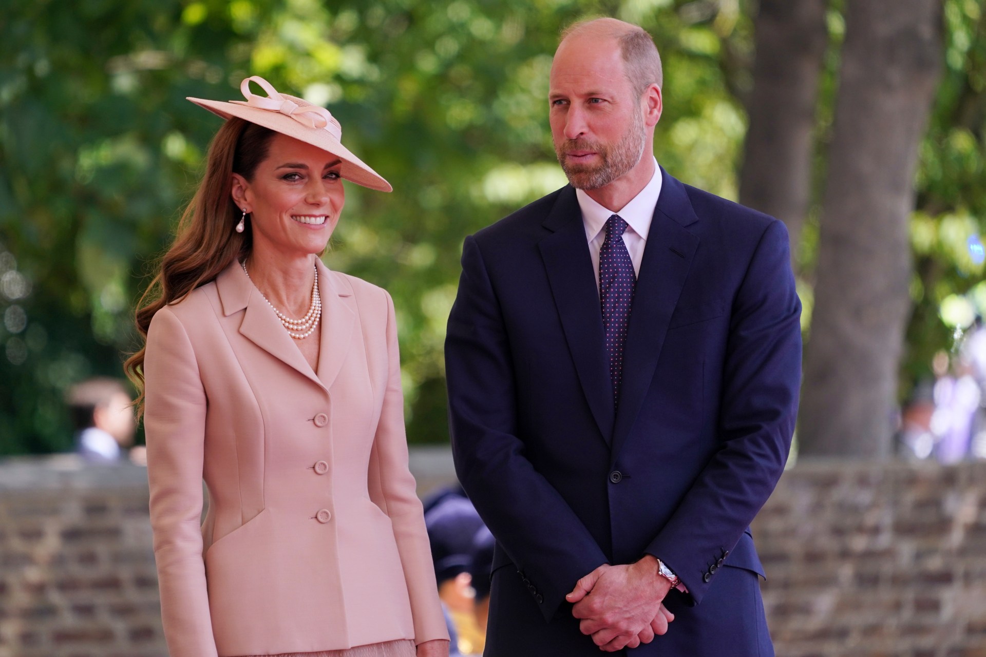 Princess of Wales in a pink suit and hat smiling next to Prince William in a navy suit at Windsor Castle.
