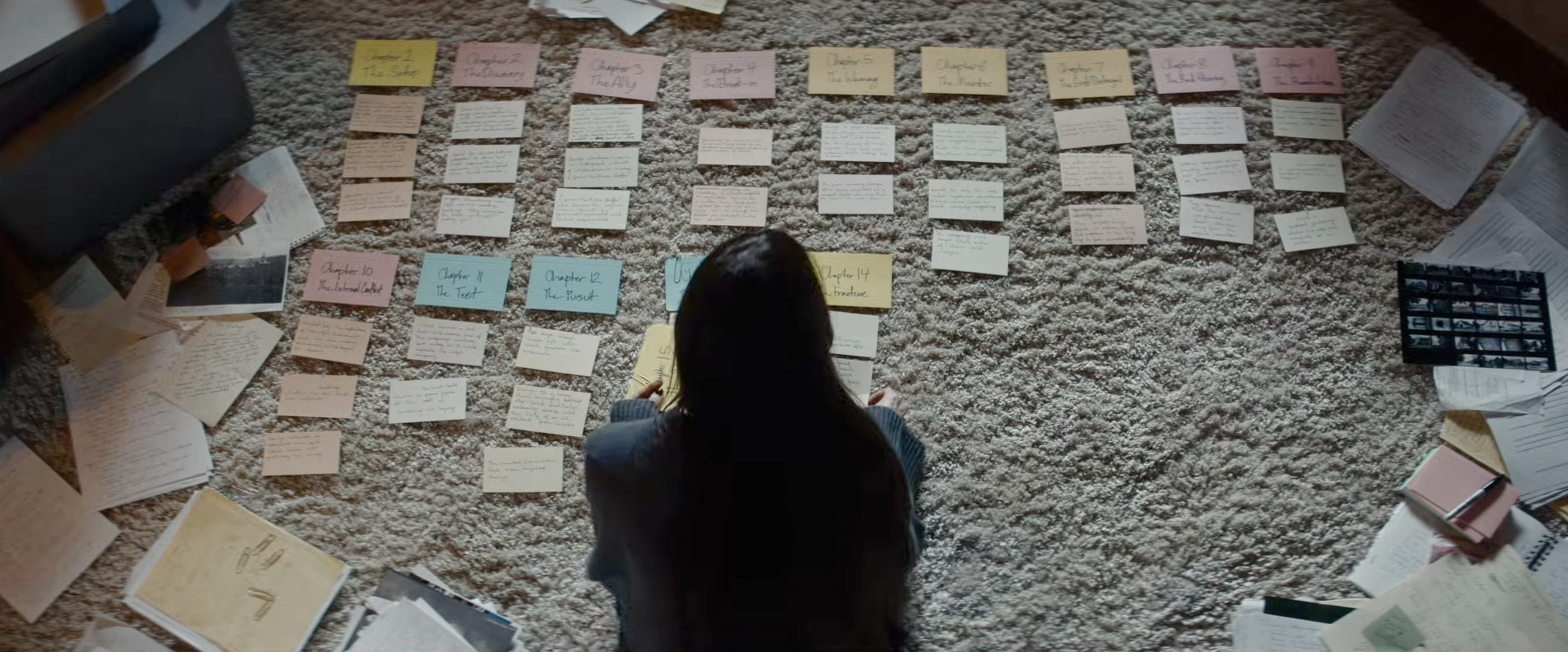 Overhead view of a woman sitting on carpet surrounded by color-coded handwritten index cards arranged by chapter, with notebooks and papers spread around her.