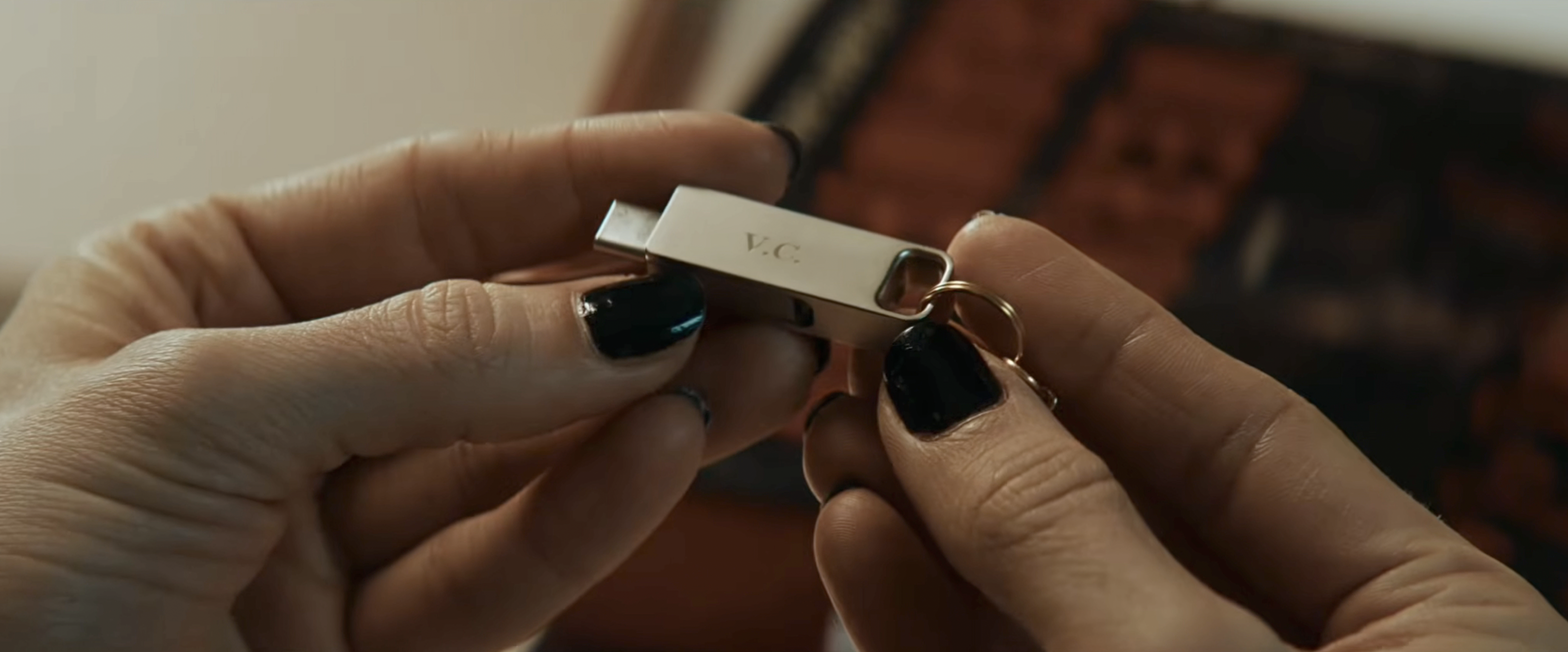 Close-up of two hands with black nail polish holding a silver USB drive engraved with the initials V.C., with a gold keyring attached.