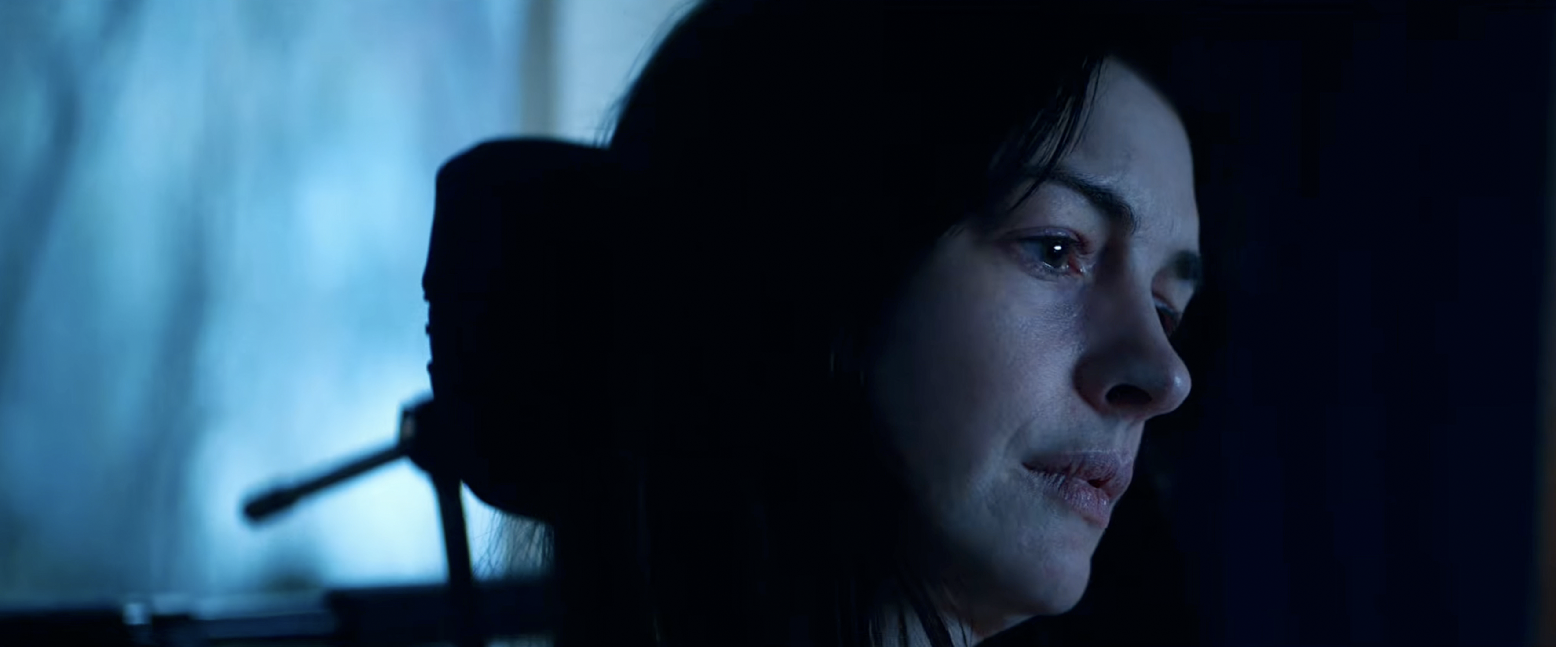 Close-up profile of a dark-haired woman illuminated by dim blue light, gazing off to the side with a pensive expression.