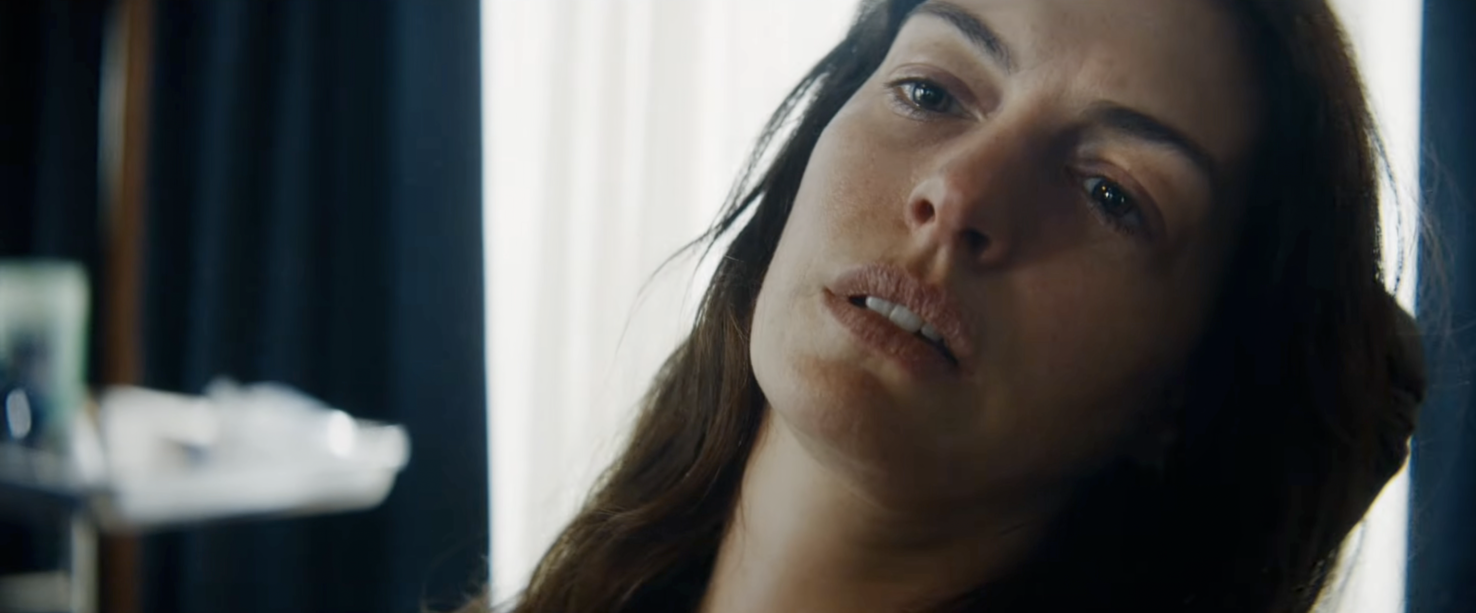 Close-up of a young woman with long dark hair looking upward with a pensive expression, soft light behind her.