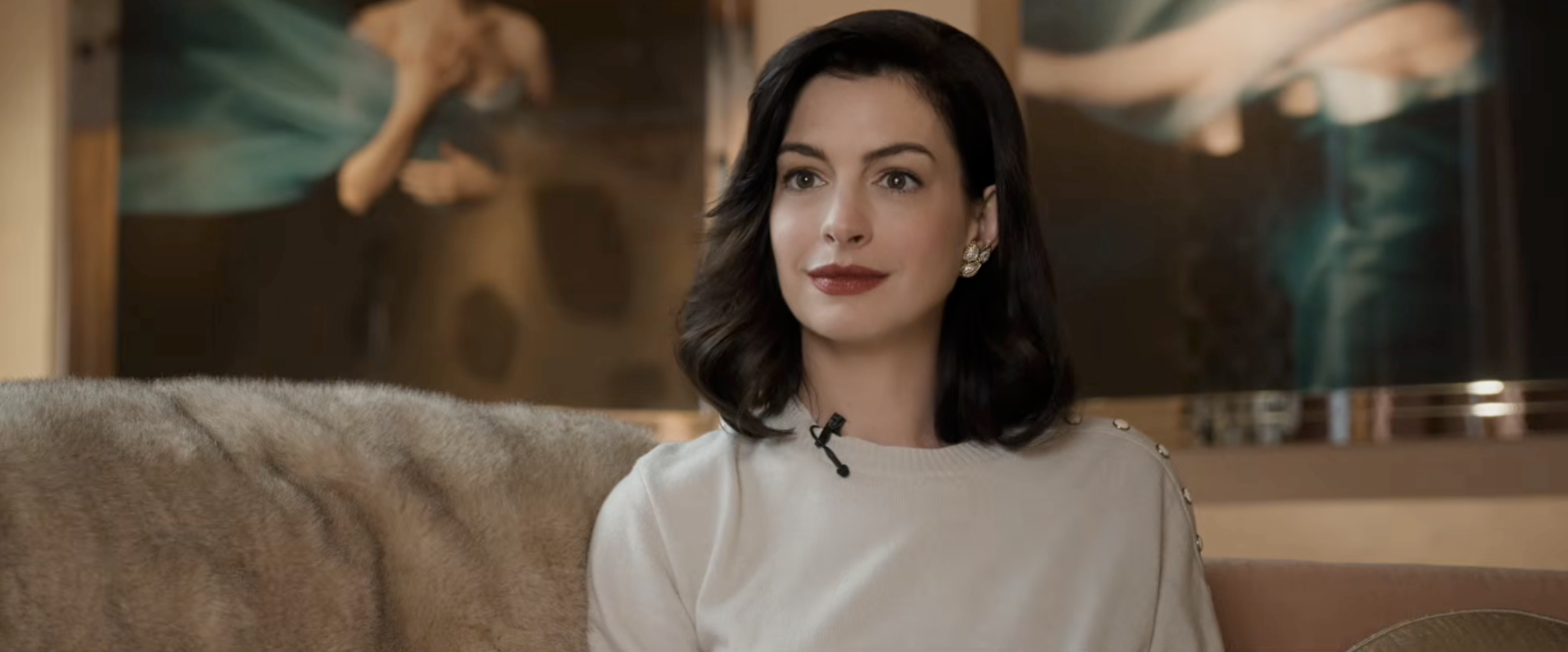 Dark-haired woman in a white top and gold earrings seated on a couch, large figurative painting visible behind her.