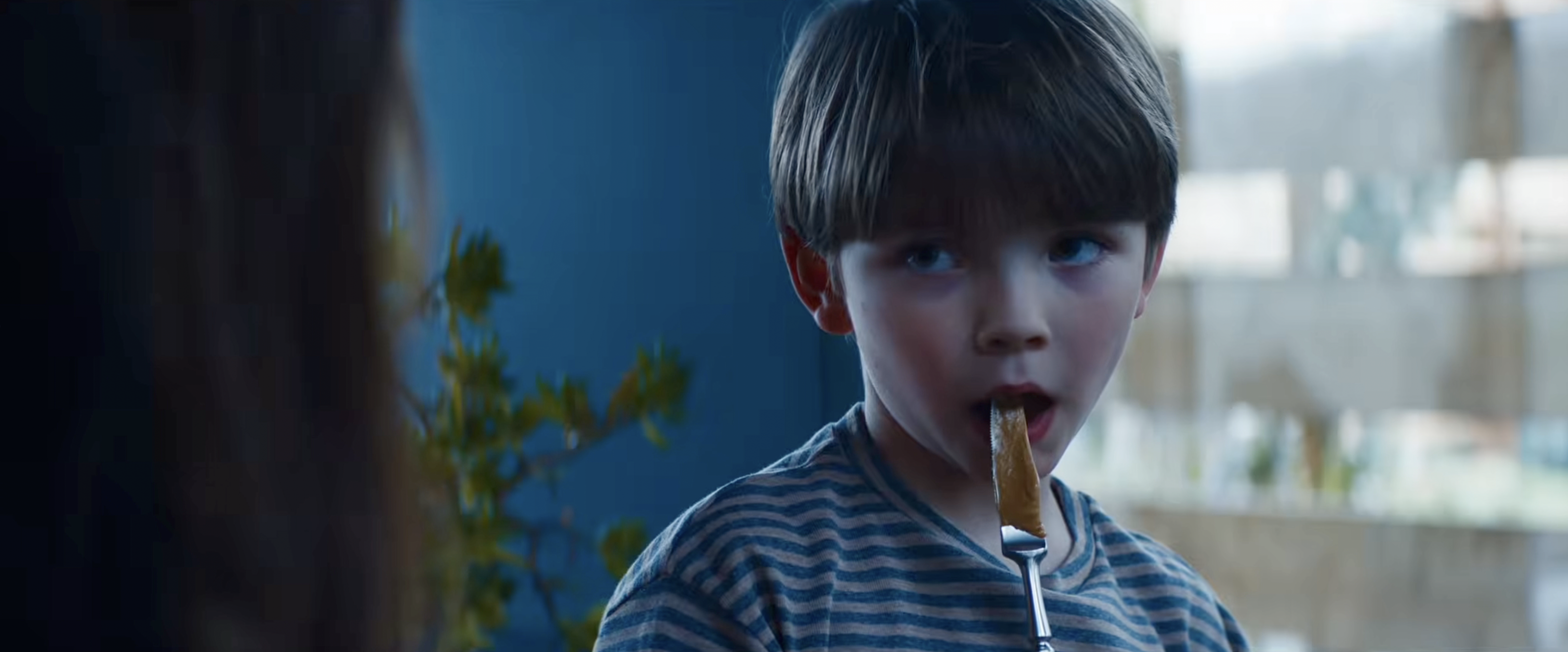 Young boy in a blue striped shirt eating food off a fork, looking sideways with mouth open.