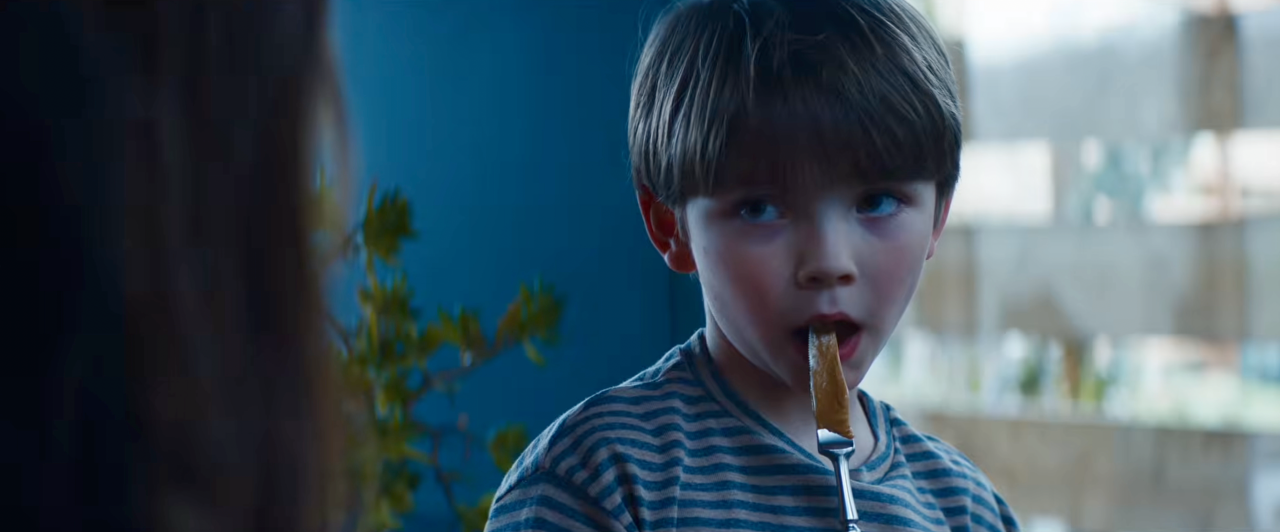 Young boy in a blue striped shirt eating food off a fork, looking sideways with mouth open.