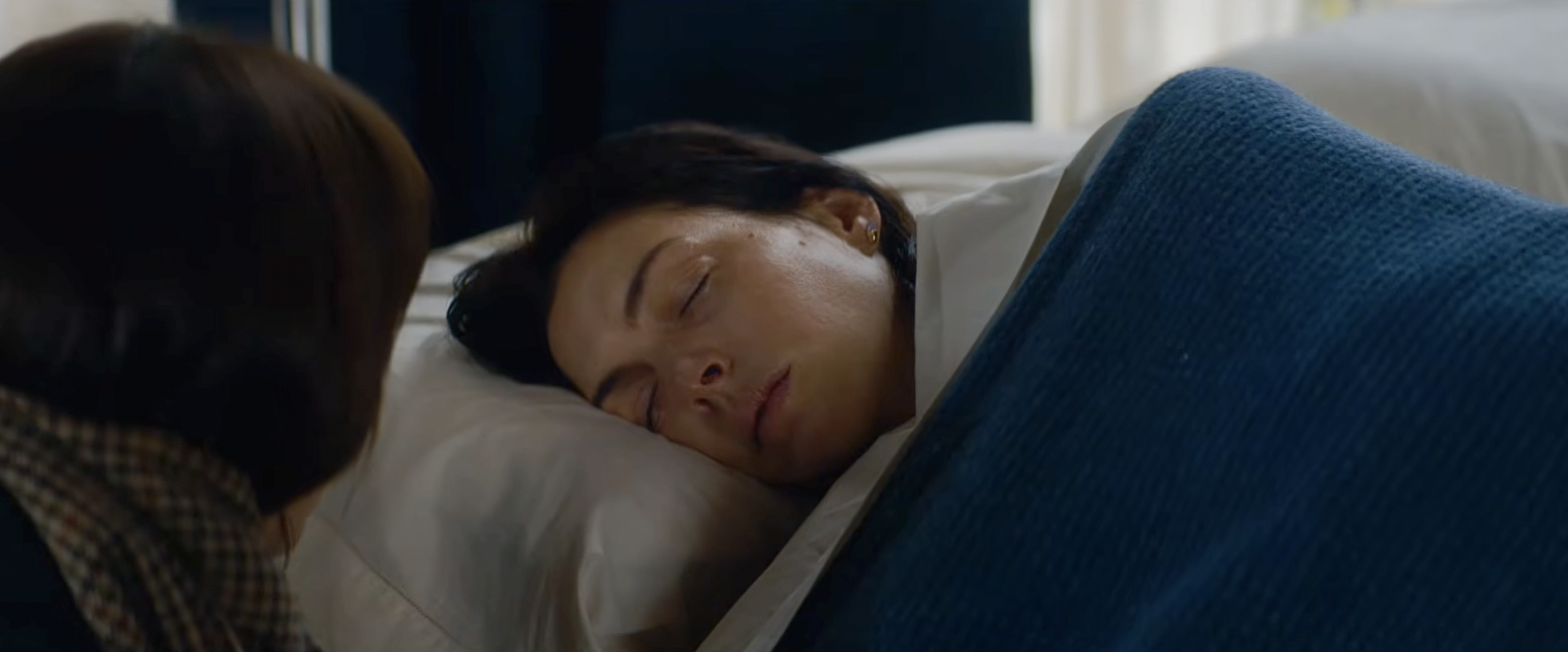 Woman with dark hair lying on a white pillow in a hospital bed, eyes closed, with another person leaning over her.