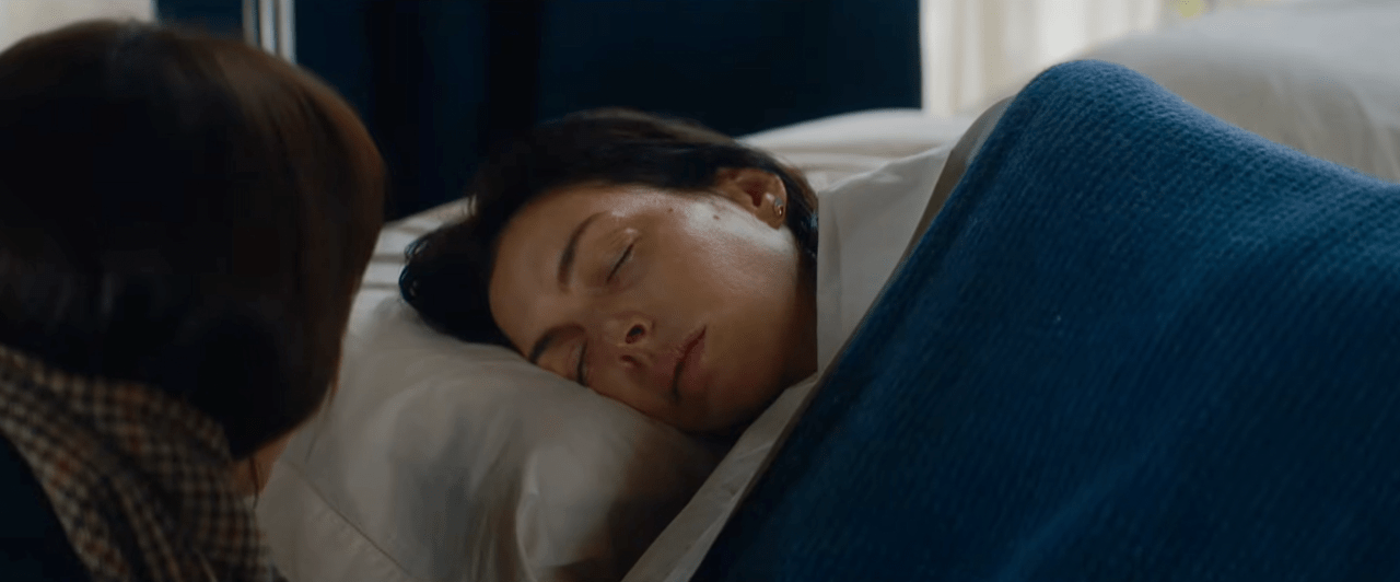 Woman with dark hair lying on a white pillow in a hospital bed, eyes closed, with another person leaning over her.