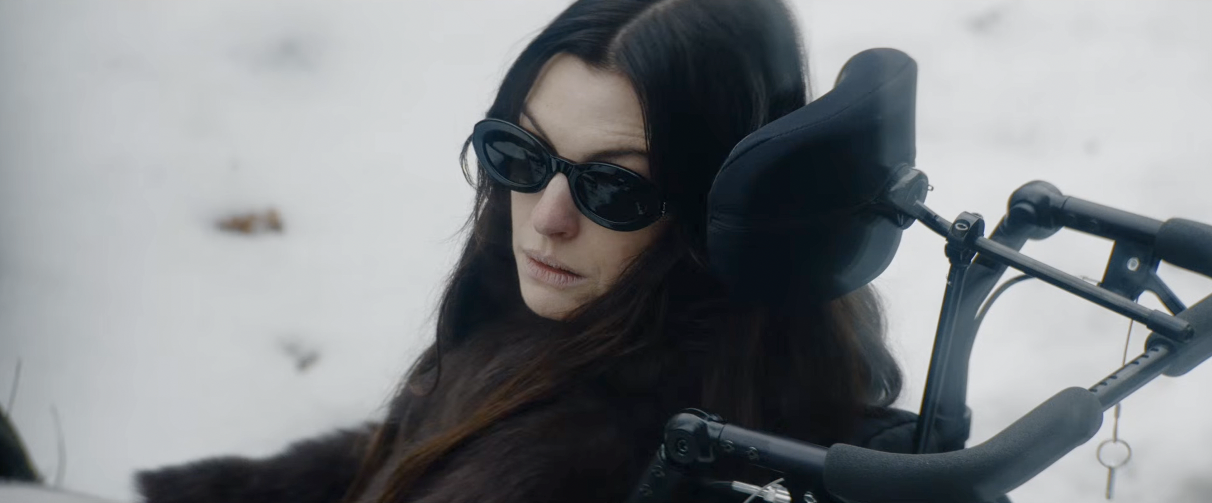 Woman with long dark hair wearing black oval sunglasses seated in a power wheelchair against a snowy background.