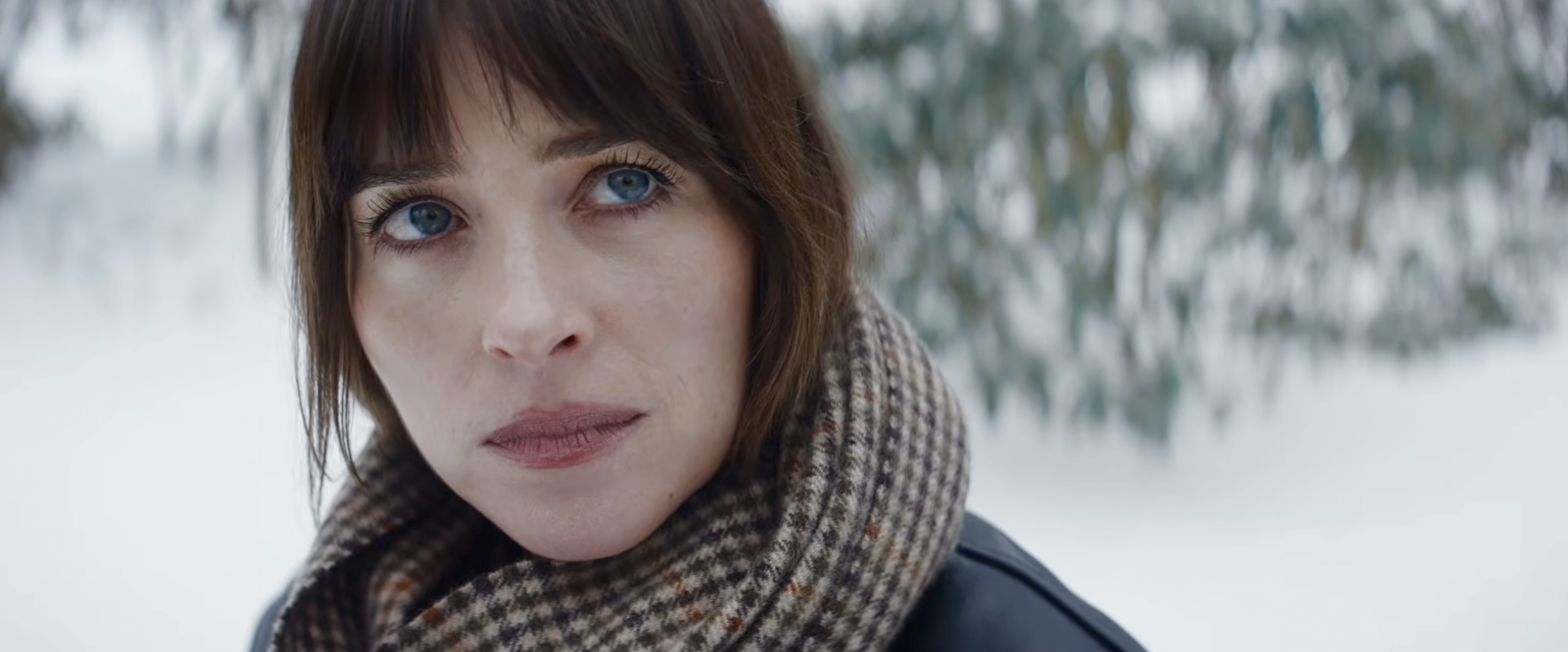 Close-up of a dark-haired woman with blue eyes wearing a plaid scarf outdoors in a snowy setting.