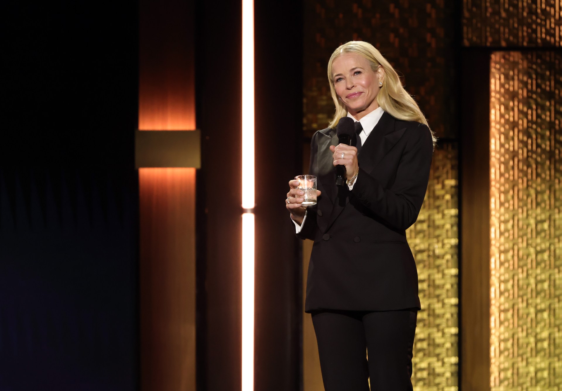 Blonde woman in a black suit holding a microphone and a glass tumbler onstage before a gold-paneled backdrop with warm lighting.