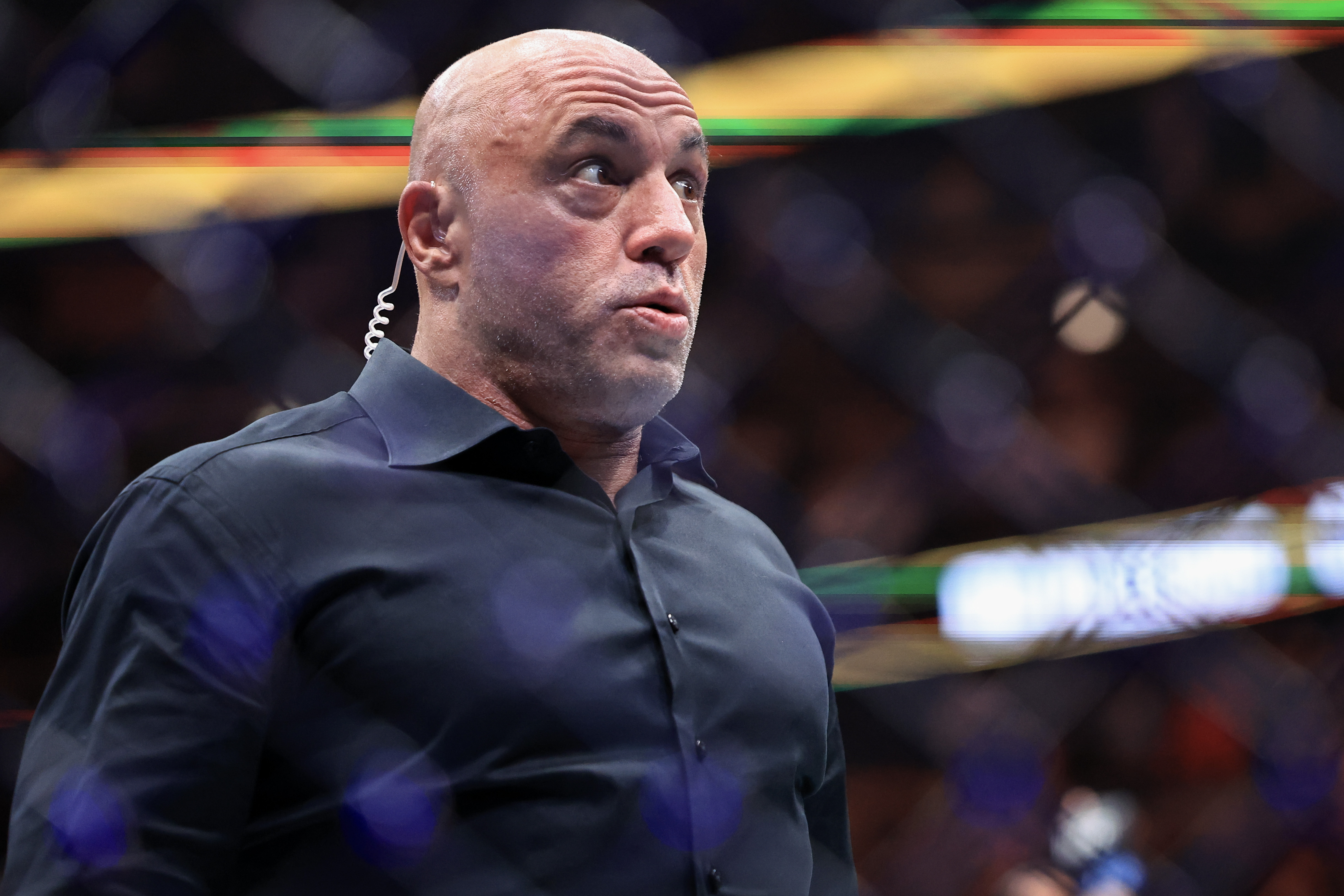 Bald man in a dark button-up shirt with a coiled earpiece, gazing upward inside a brightly lit sports arena.
