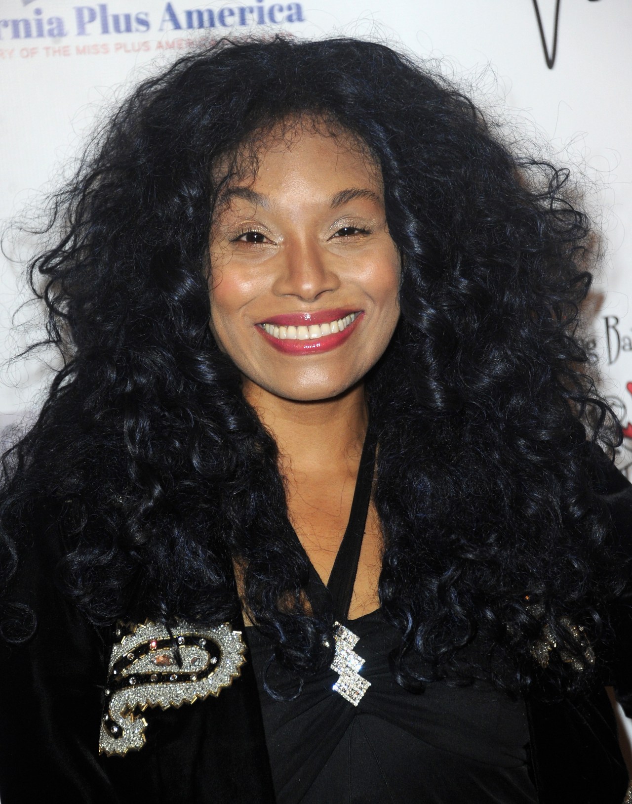 Actress Christy Knowings smiling at a red carpet event, wearing a black embellished jacket with voluminous curly dark hair.