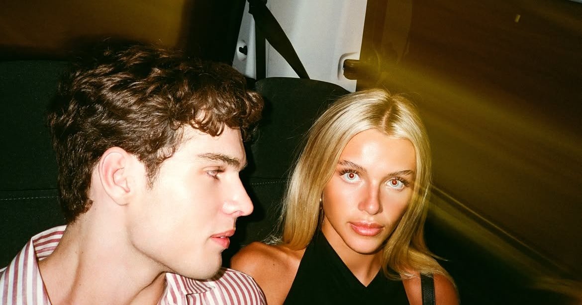 A young man with curly brown hair and a young blonde woman sitting together in the backseat of a car, film photo with lens flare.