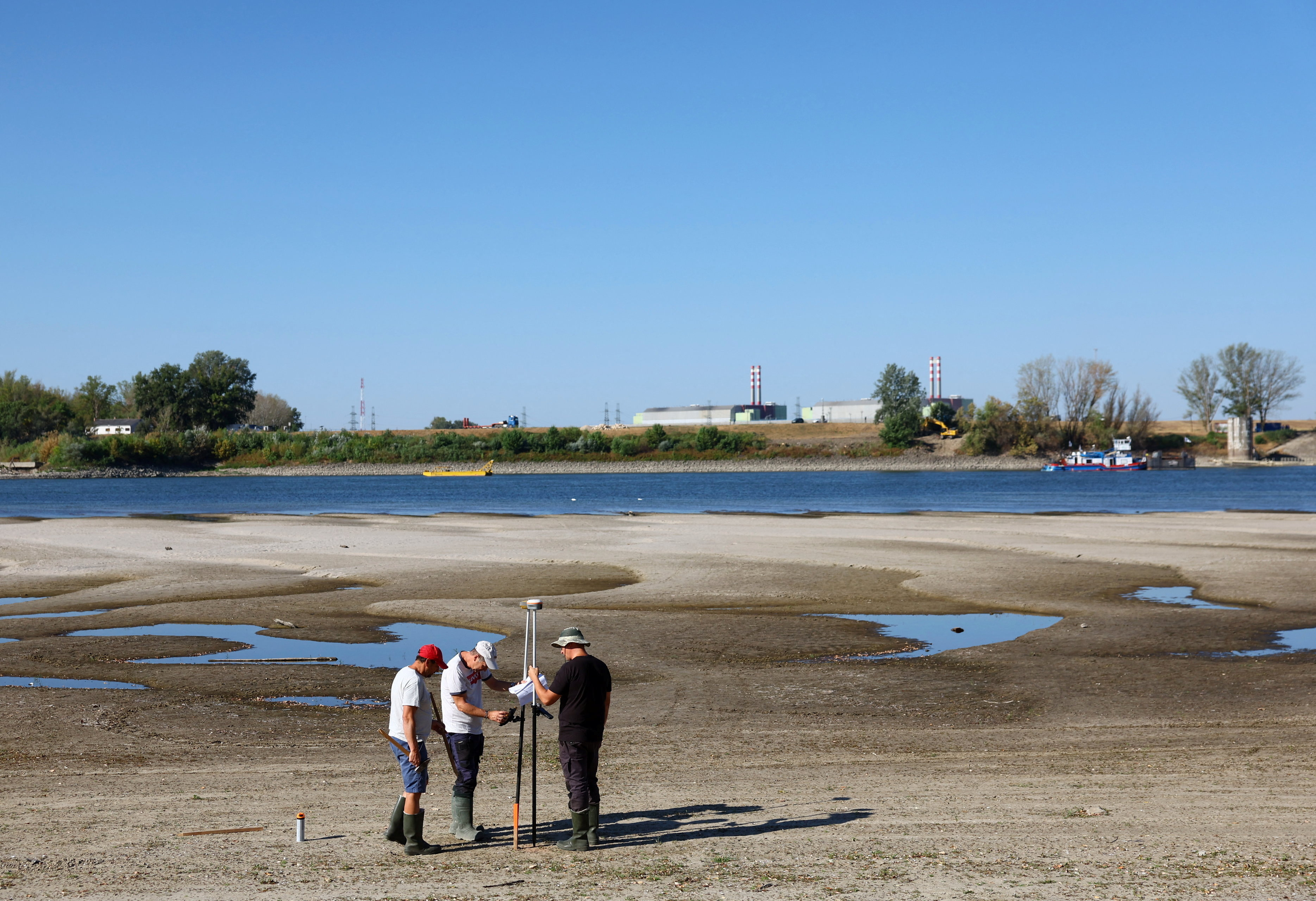 Three workers in rubber boots huddle around a surveying tripod on a wide, exposed sandy riverbed, with shallow water channels and industrial smokestacks visible in the background.