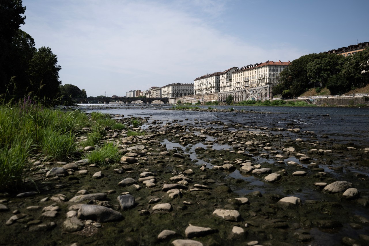 Rocky, partially dried riverbed of the Po River in Turin, with shallow water and exposed stones in the foreground and historic buildings along the far bank.
