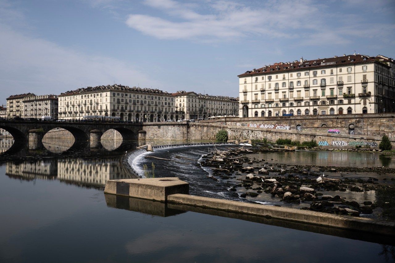 Wide-angle view of the low Po River in Turin, showing a stone arch bridge reflected in shallow water, with exposed rocks and graffiti-covered walls along the riverbank.