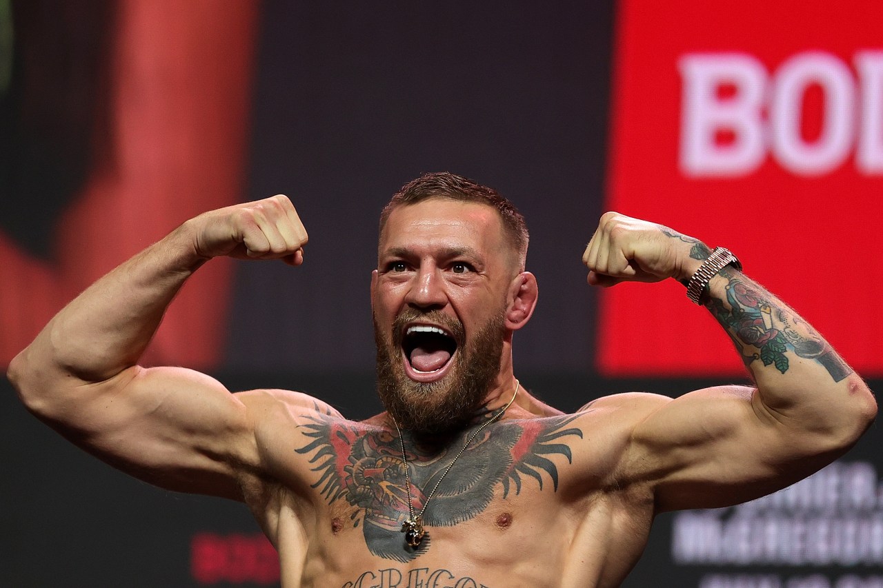 Shirtless muscular man with beard and chest tattoos flexing both arms overhead with mouth open, in front of a red event backdrop.