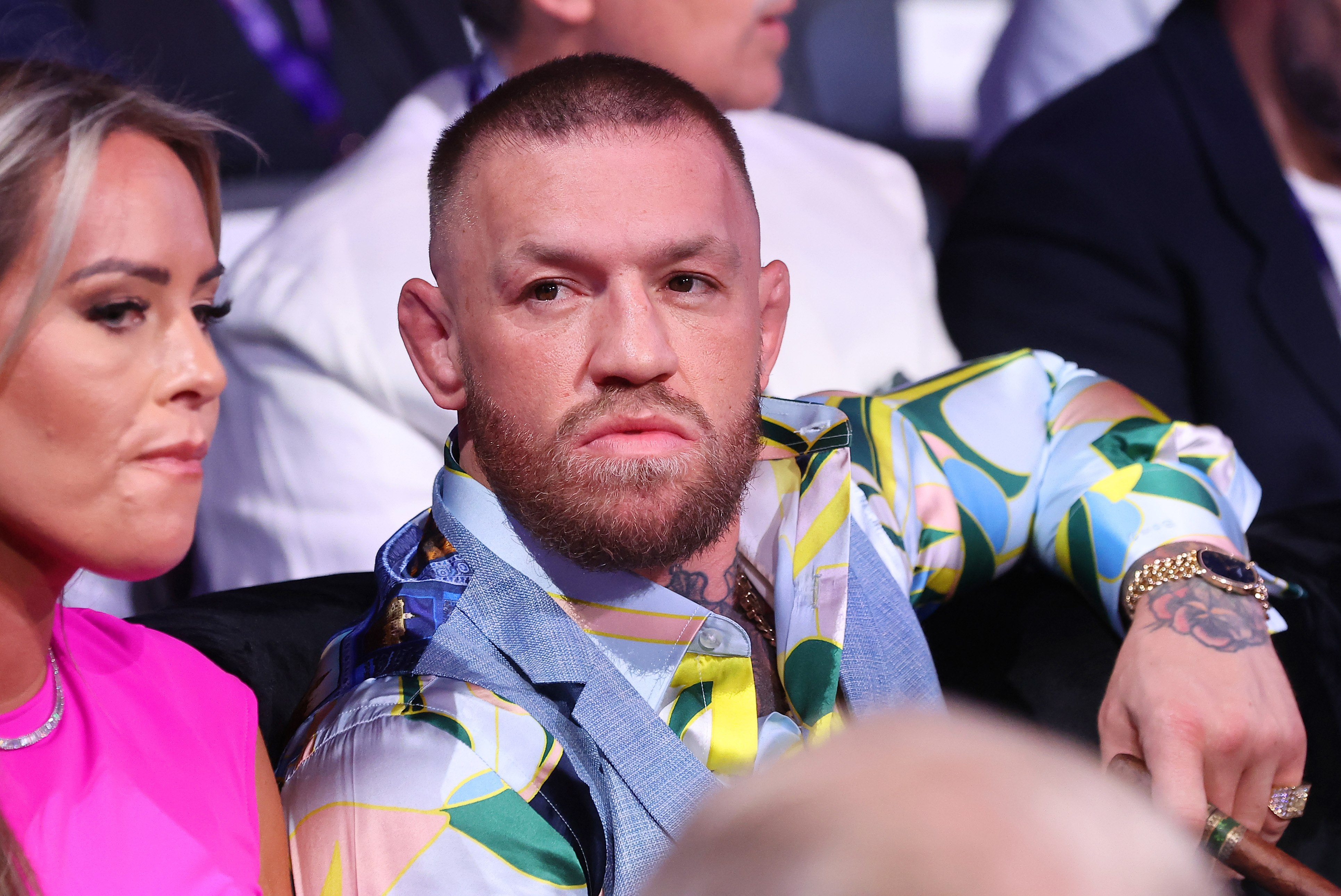 Short-haired bearded man in a blue and green patterned shirt seated in a crowded arena, looking straight ahead with a serious expression.