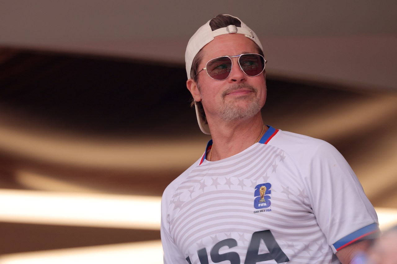 Man in a white FIFA World Cup 2026 USA jersey and a backwards cap wearing aviator sunglasses, looking upward in a stadium.