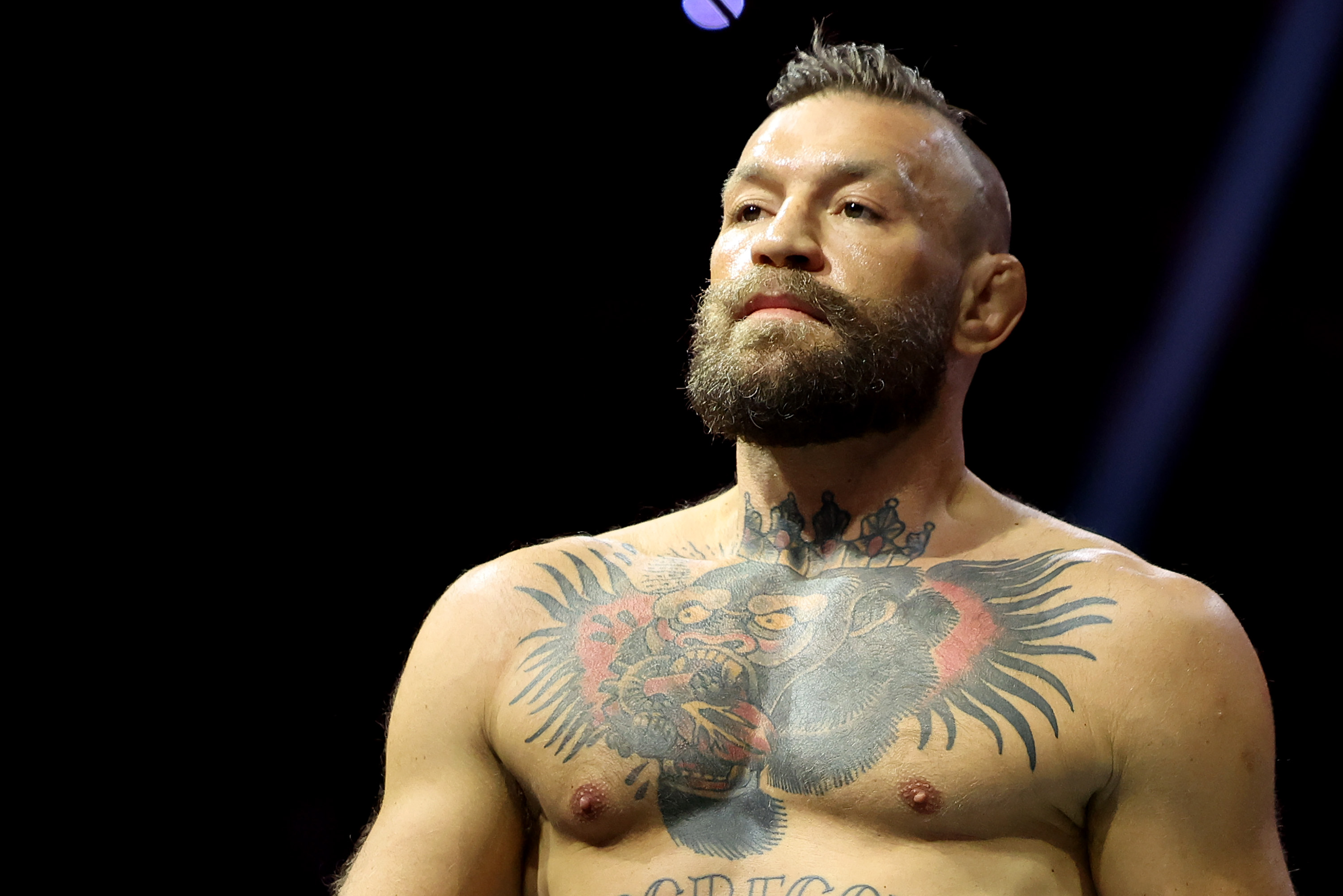 Shirtless man with a large gorilla chest tattoo and short beard gazes forward under arena lights against a black background.