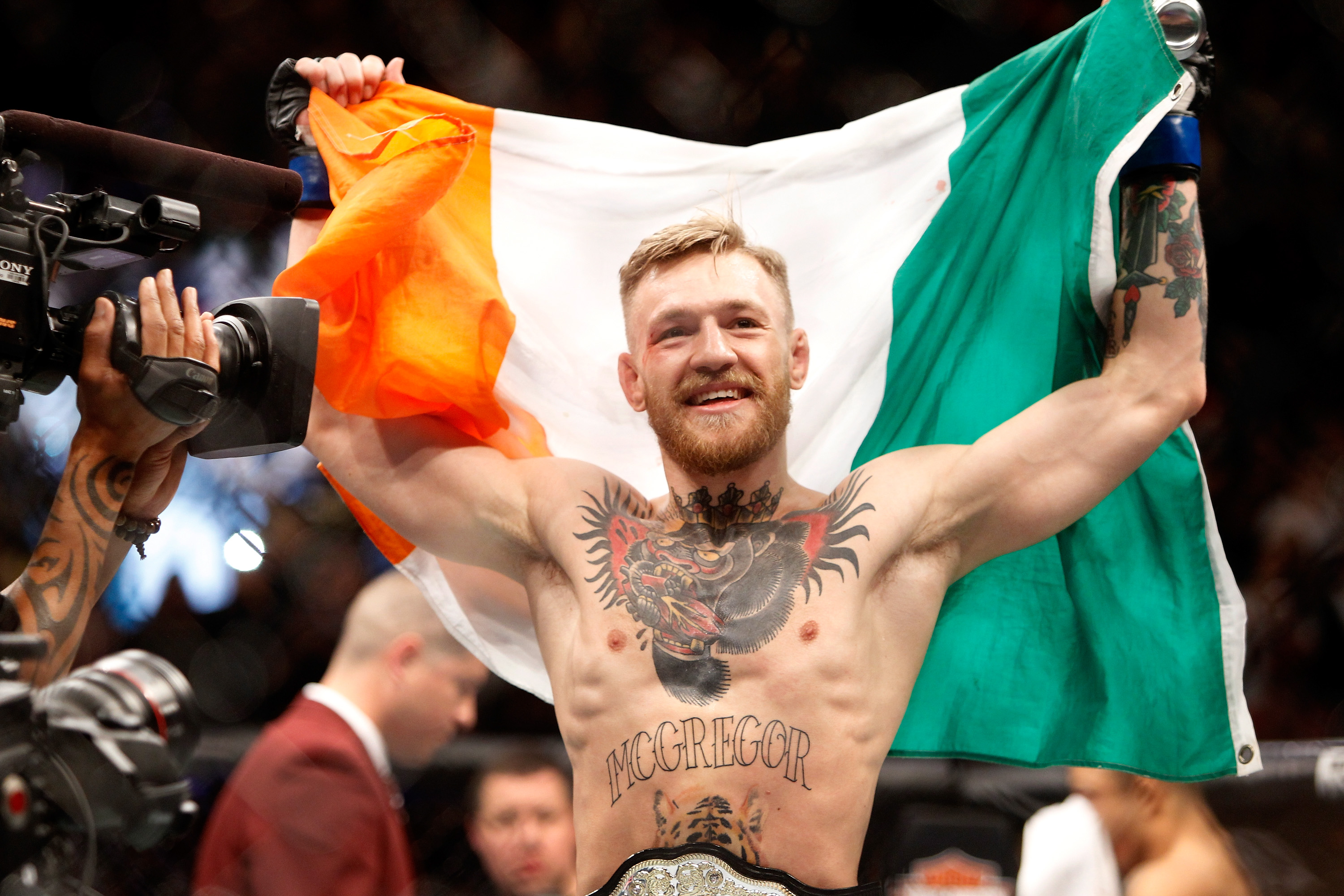 Shirtless man with red beard holding an Irish flag aloft with both arms outstretched, a UFC championship belt visible at his waist.