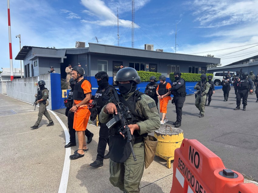 Armed officers in black tactical gear and helmets escort two handcuffed men in orange jumpsuits across an airport tarmac in Costa Rica.