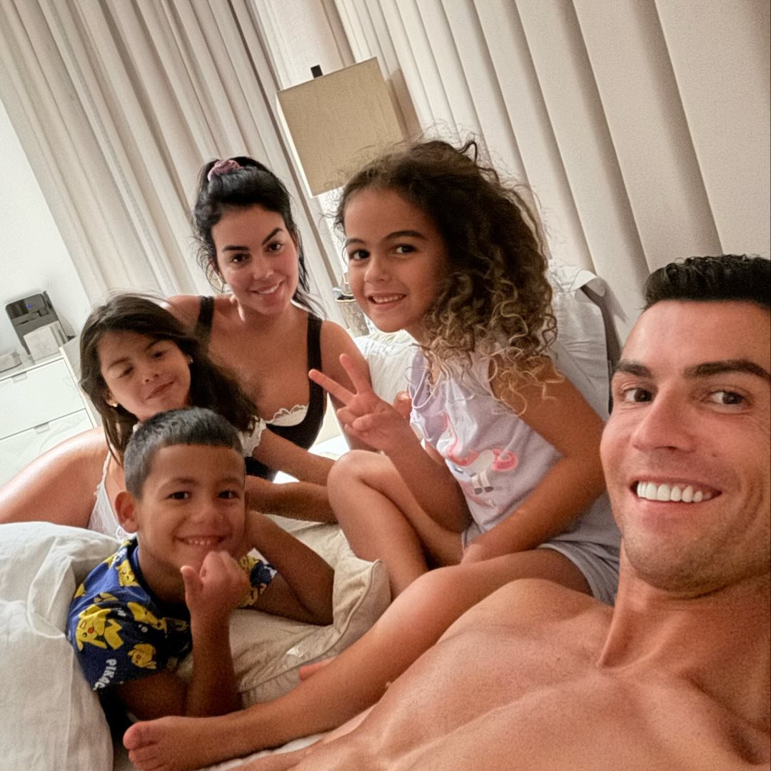 Man takes a bedroom selfie with a woman and three young children lounging on a white bed.