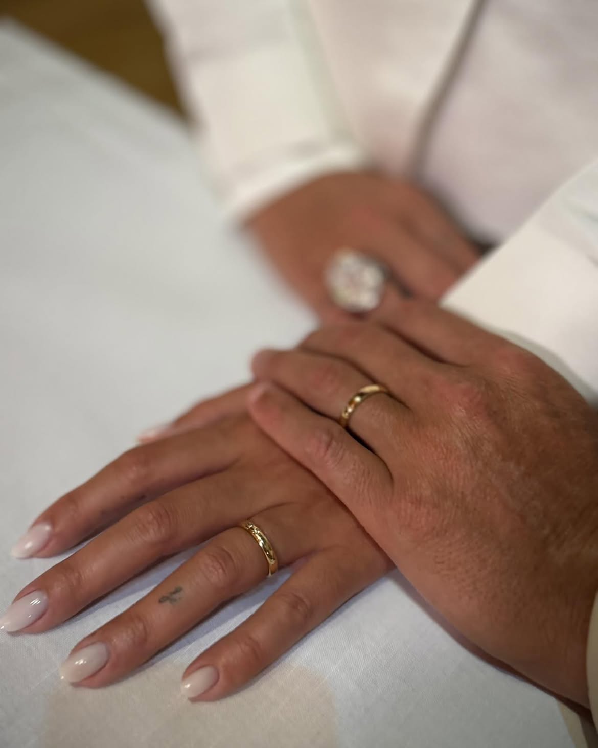 Close-up of two hands wearing matching gold wedding bands, one hand with a large oval diamond engagement ring and a small cross tattoo.