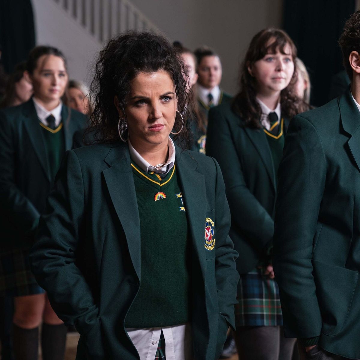 A curly-haired girl in a green school uniform with hoop earrings and a rainbow badge stands with hands behind her back among other students in a hall.