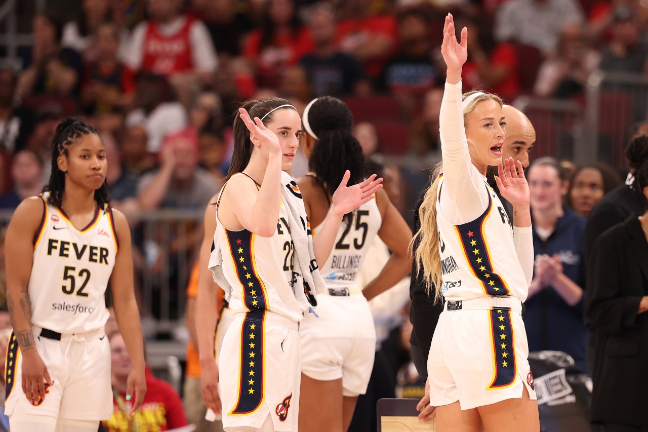 Indiana Fever players Caitlin Clark and Sophie Cunningham raising their hands and waving on the sideline alongside teammates.